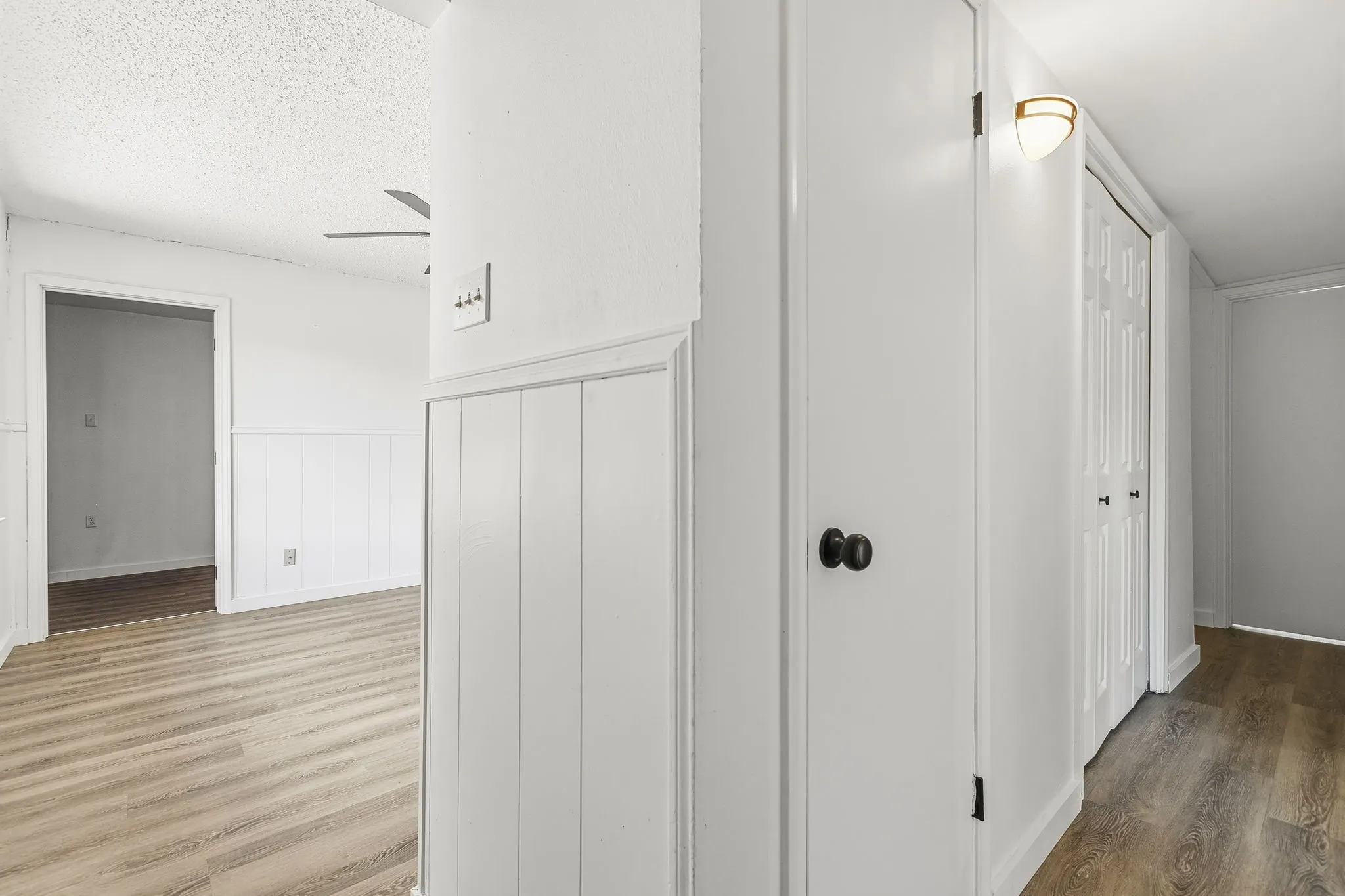 Wood-finish flooring, white paneled accent wall, matte black door hardware, ceiling fan, and wall sconce lighting
