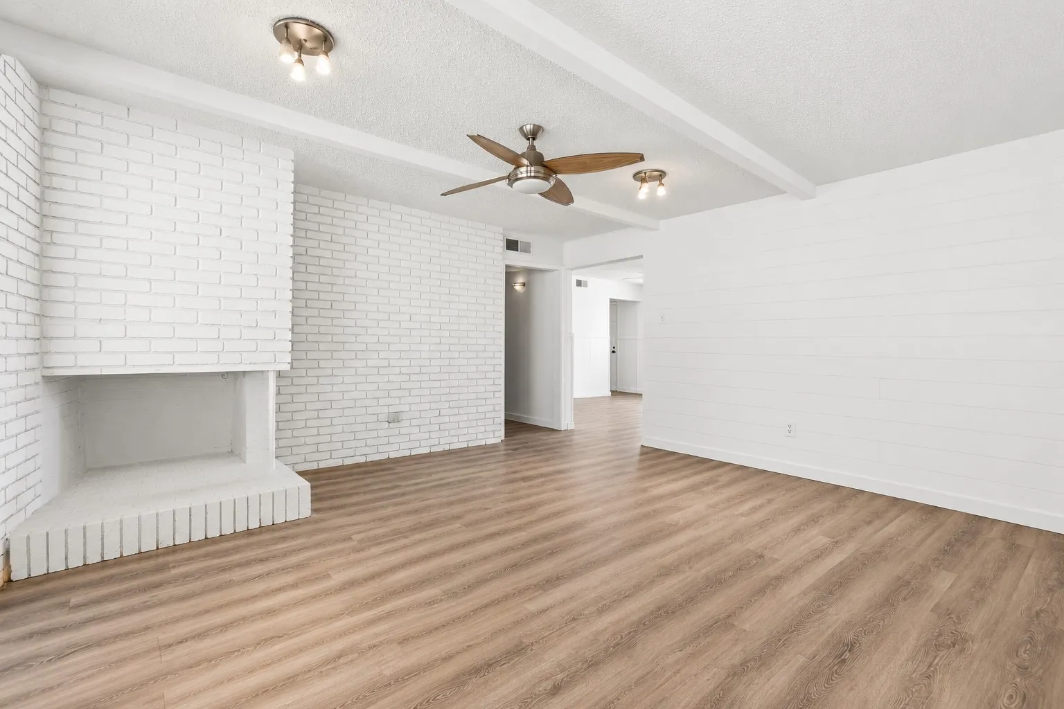 Wood-finish flooring, white brick fireplace, white shiplap accent wall, exposed ceiling beams, and ceiling fan with integrated lighting