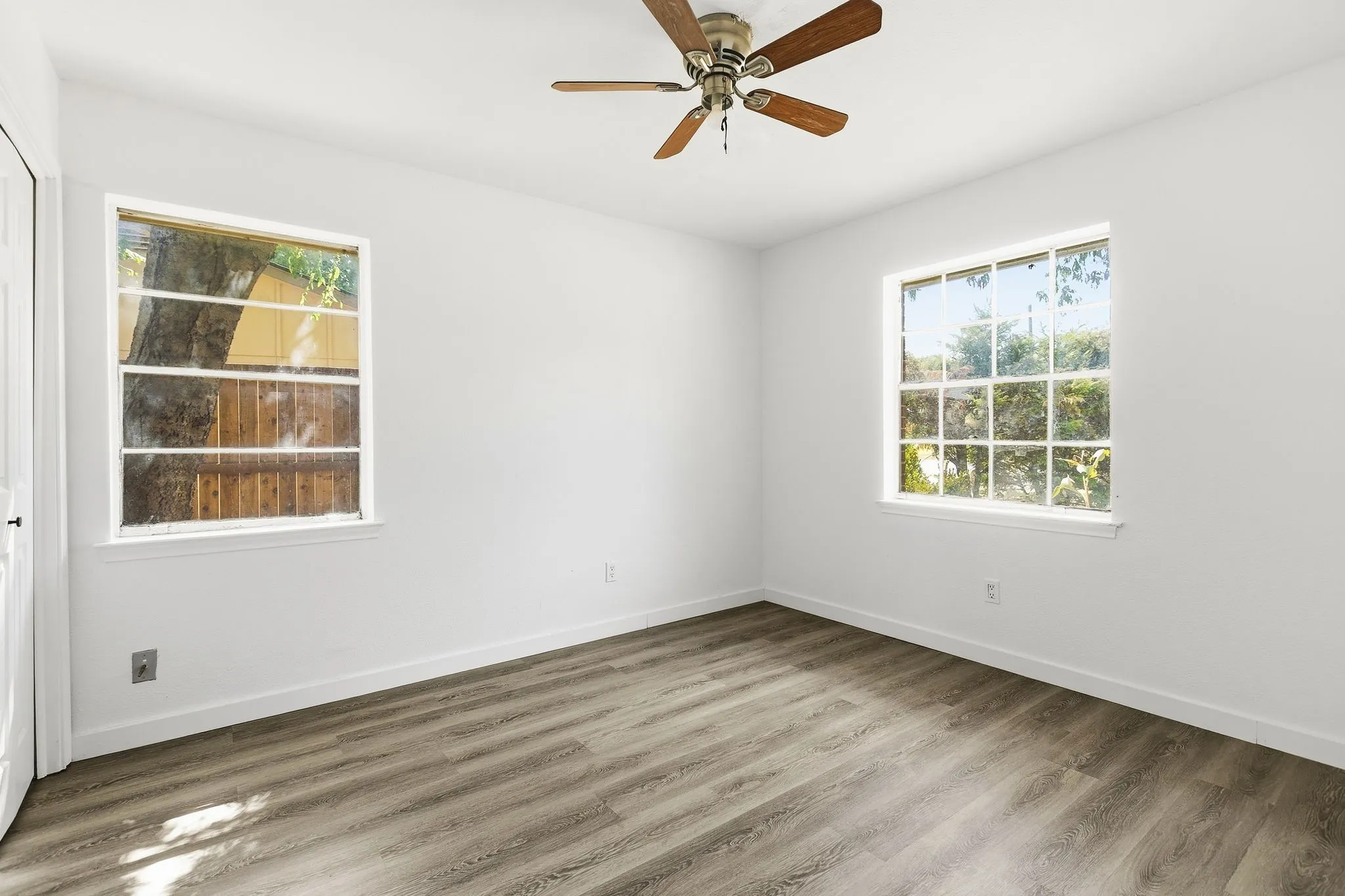 Room featuring wood-finish flooring, two windows with grids, white painted walls, and a ceiling fan with wood-tone blades