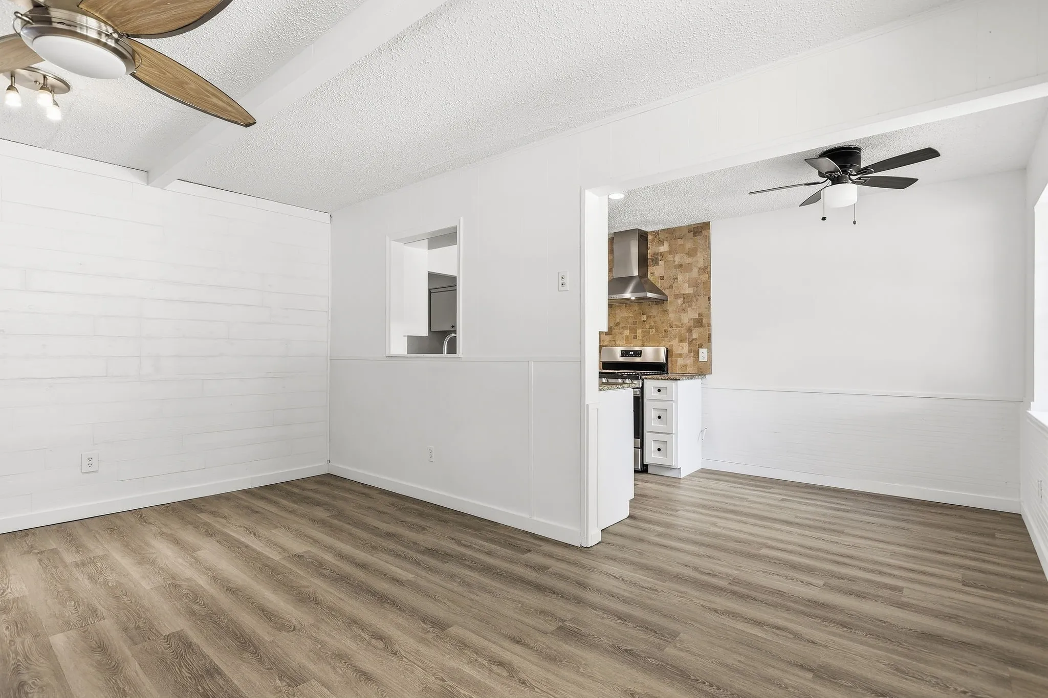 Wood-finish flooring throughout, white shiplap accent wall, two ceiling fans with light fixtures, kitchen area with stainless steel range hood, and white cabinetry