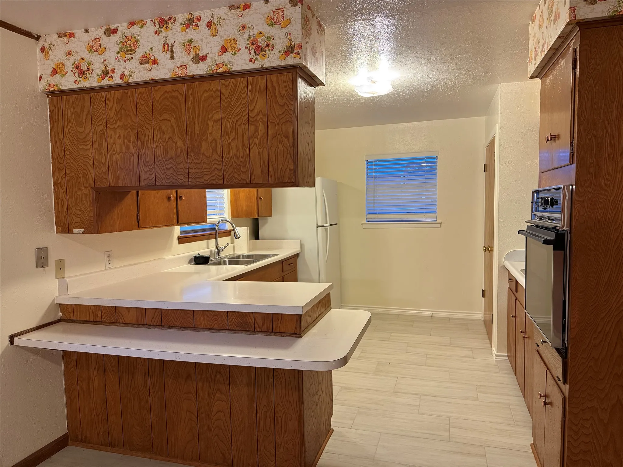 Kitchen with light countertops, a peninsula, brown cabinetry, freestanding refrigerator, and a textured ceiling