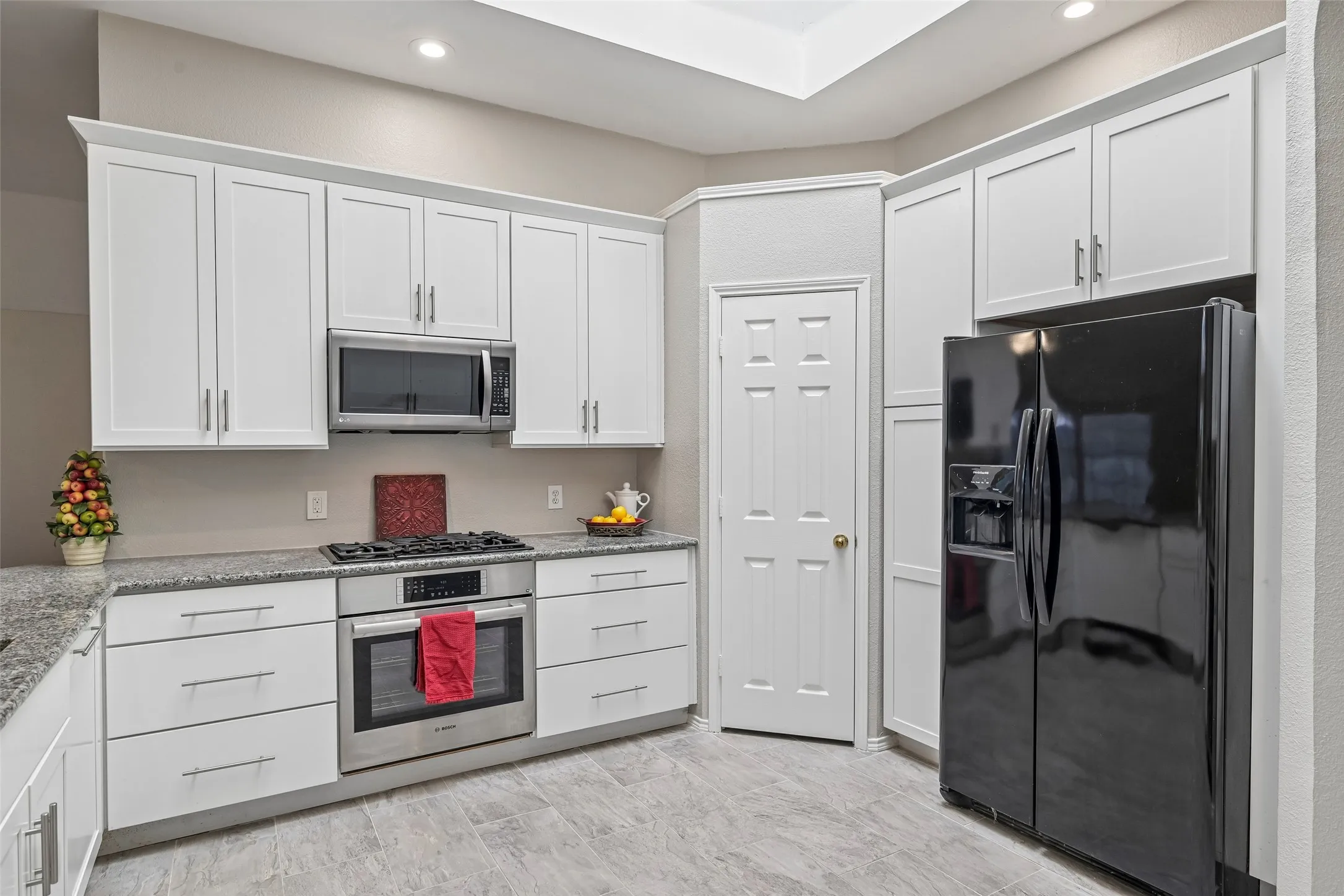 Modern kitchen featuring white shaker-style cabinetry with chrome hardware, stainless steel appliances including a gas range and black French-door refrigerator, granite countertops, recessed lighting, and neutral tile flooring throughout