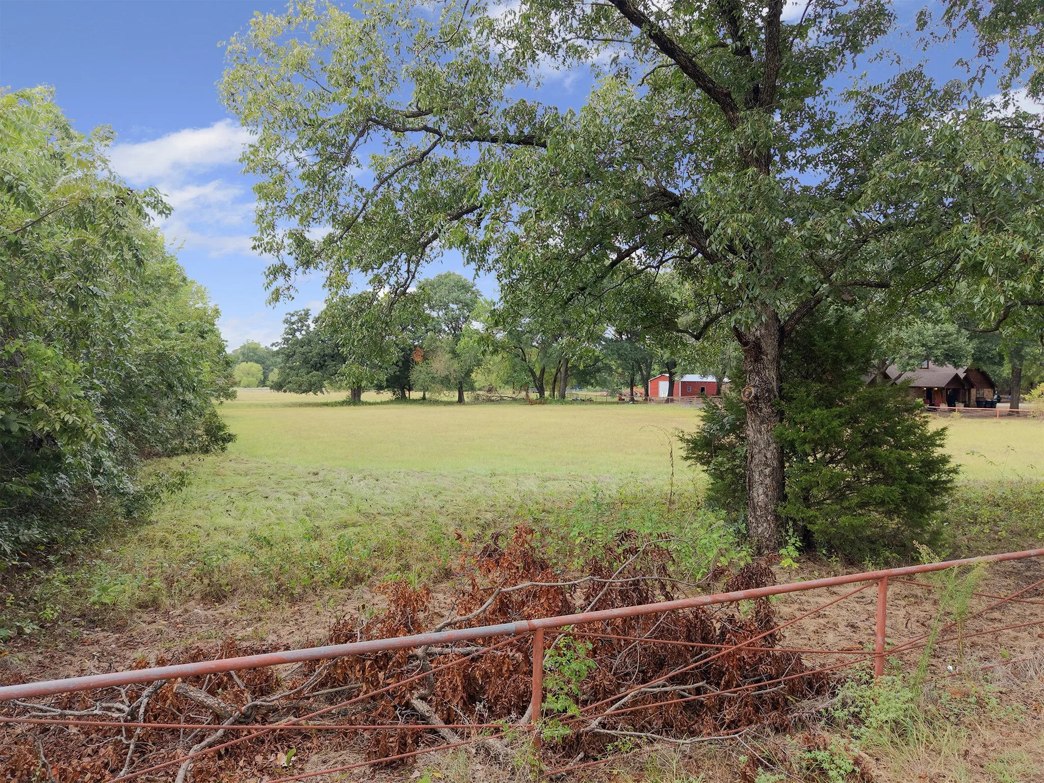 Expansive green space, mature trees, distant red barn, distant brown multi-story structure, and a metal fence