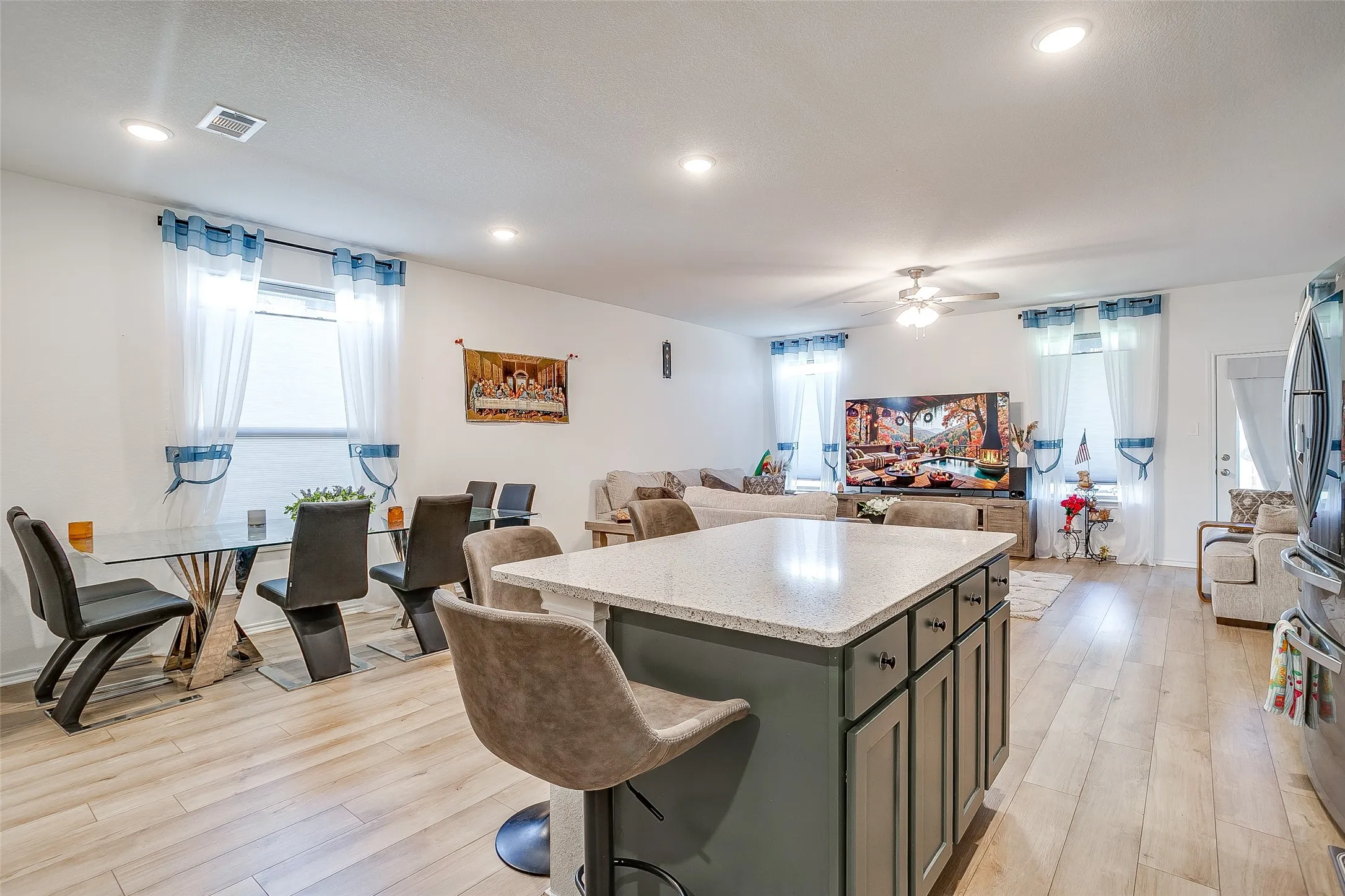 Open-concept living space featuring a large kitchen island with storage cabinetry and seating, wood-finish flooring throughout, recessed ceiling lighting, and multiple windows with blue-toned window treatments providing natural light