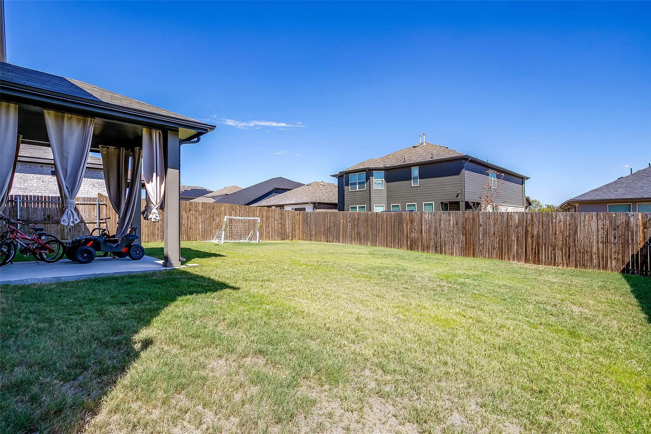Expansive backyard with a lush lawn, privacy fencing, and a covered patio featuring drapes