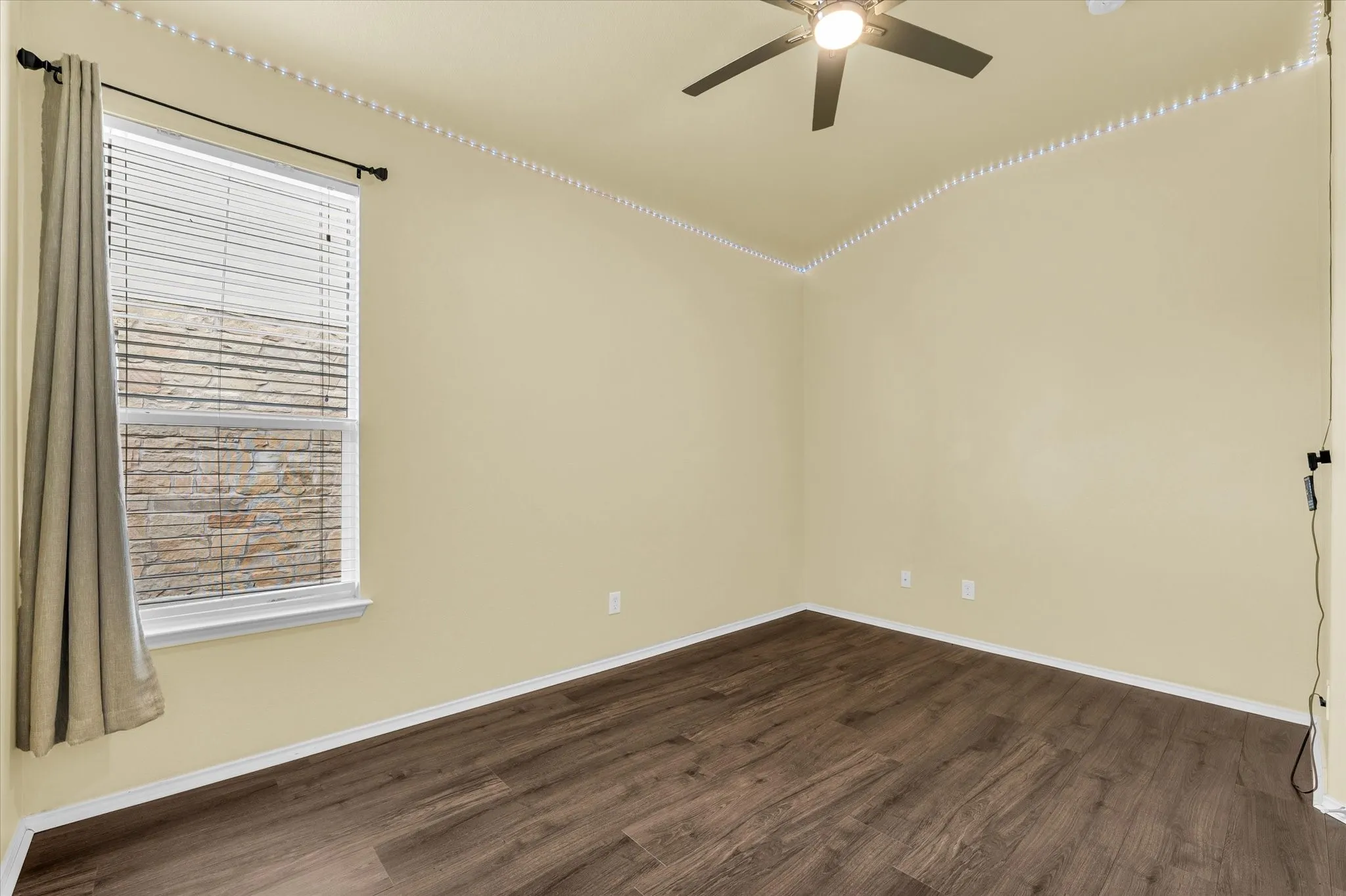 Wood-finish flooring with crisp white baseboards, a single window with blinds and a drapery rod, and a ceiling fan