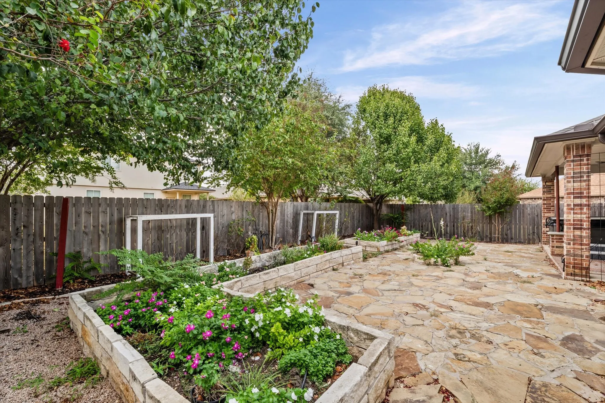 Expansive backyard featuring a natural stone patio, raised garden beds with established plantings, a wooden privacy fence, and mature trees