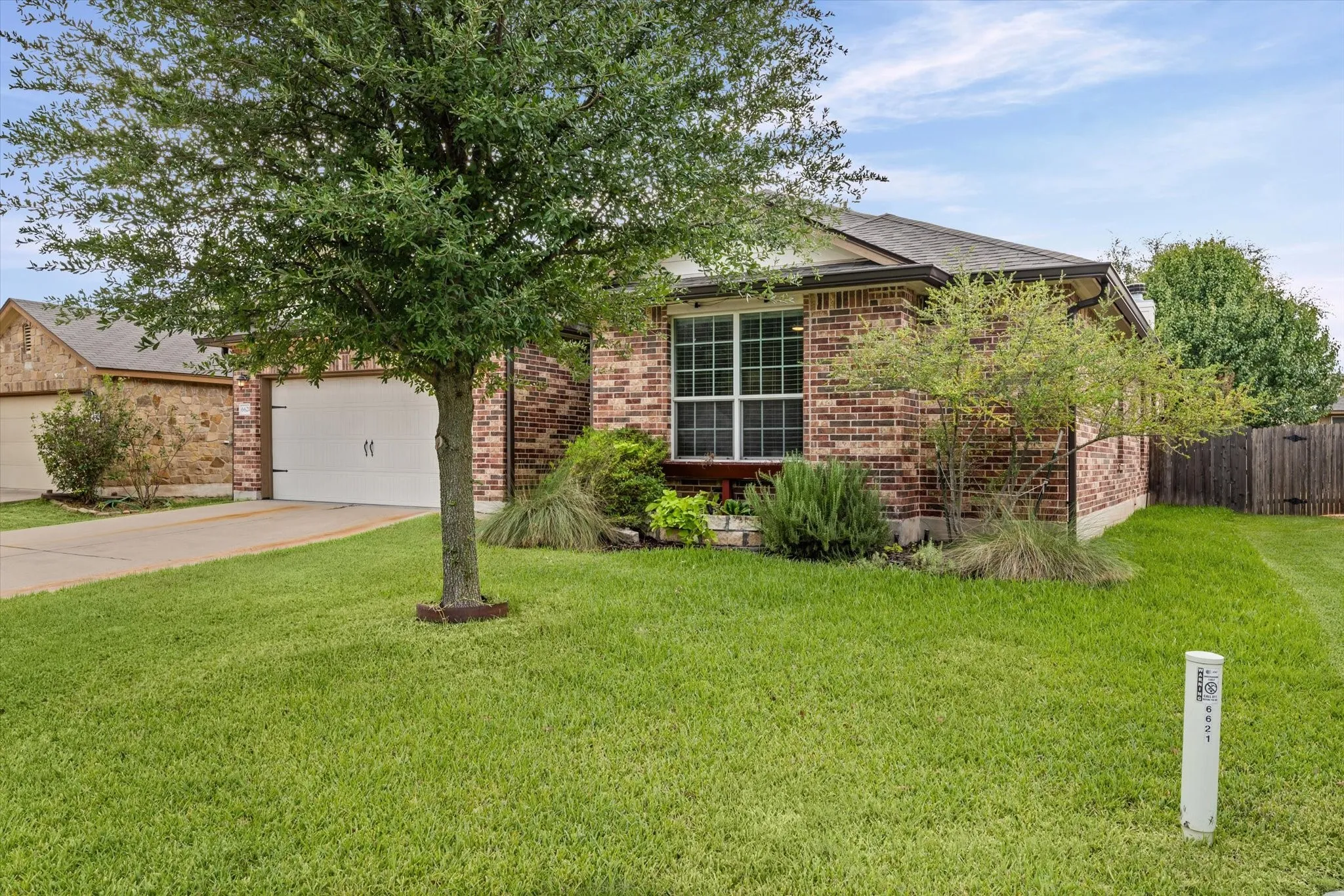 Brick exterior with a mature tree, an attached garage, a manicured lawn, and established landscaping