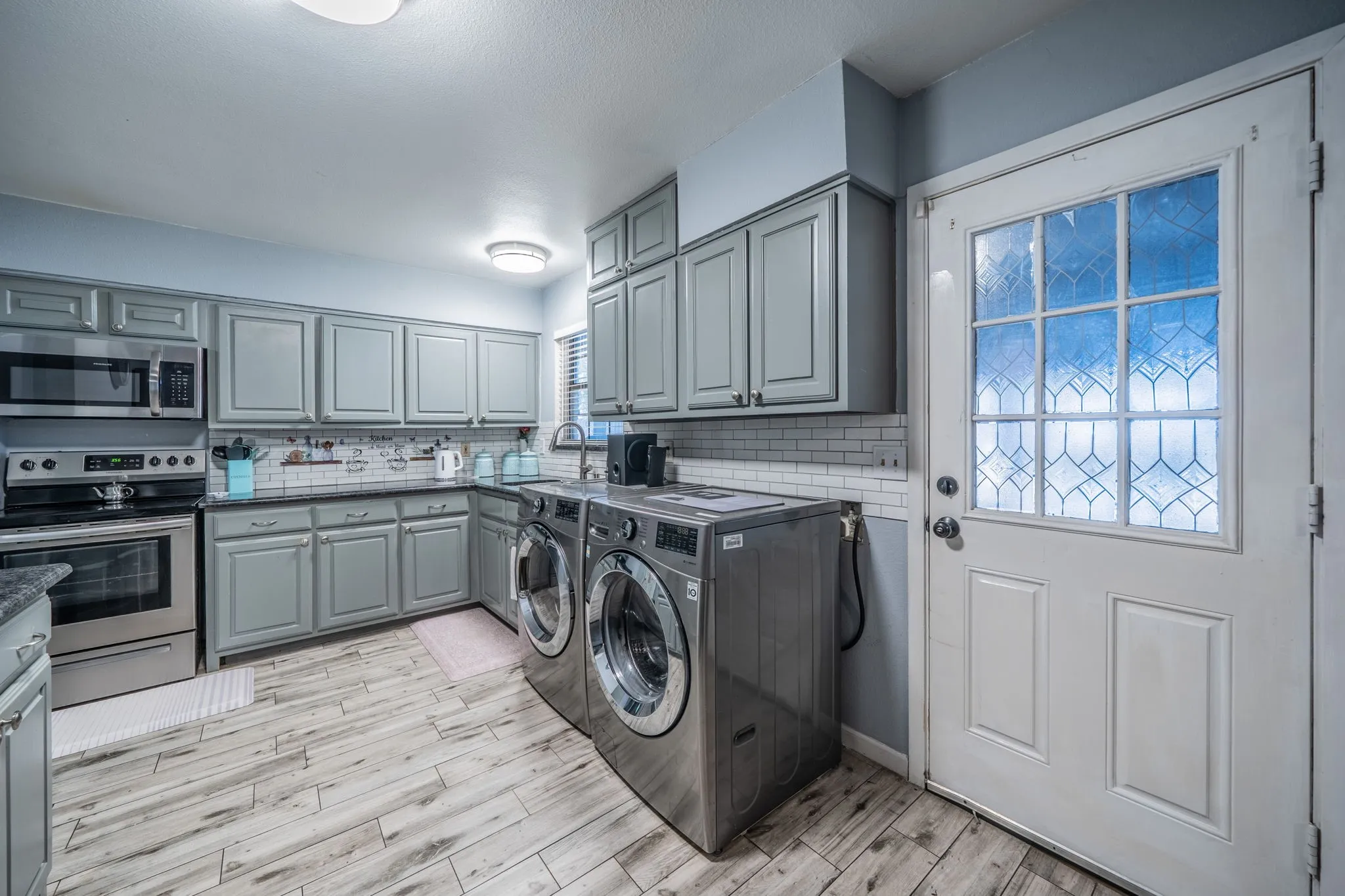 Gray cabinetry with brushed nickel hardware, subway tile backsplash, stainless steel appliances, wood-finish flooring, and a decorative glass panel door