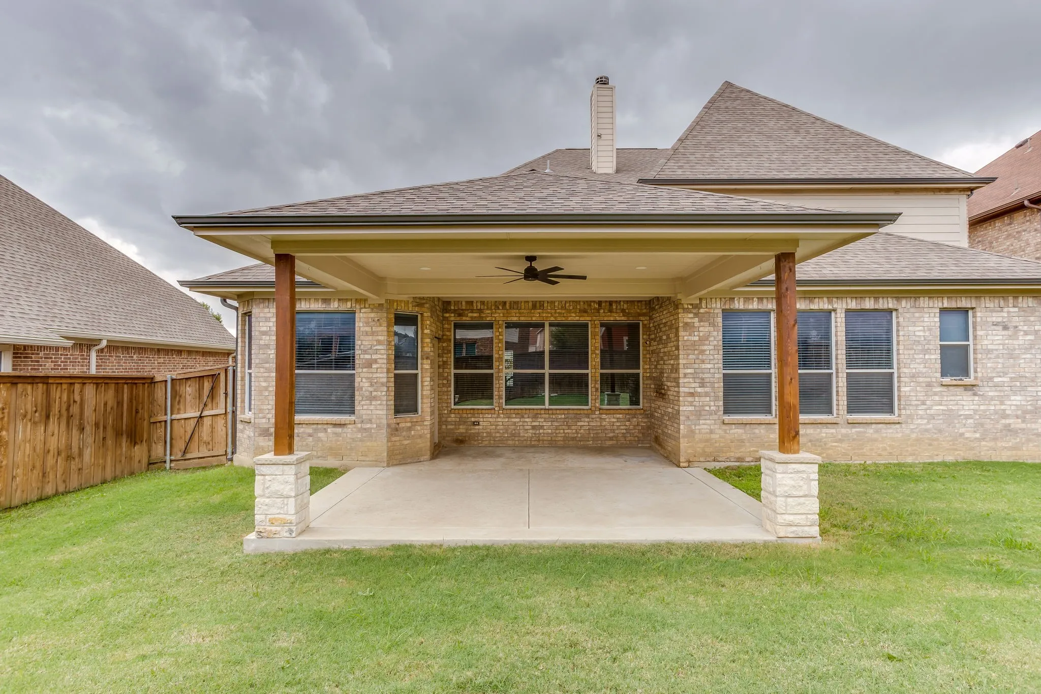 Covered concrete patio with a ceiling fan, supported by stone-clad and wood-finish posts, set against a brick exterior