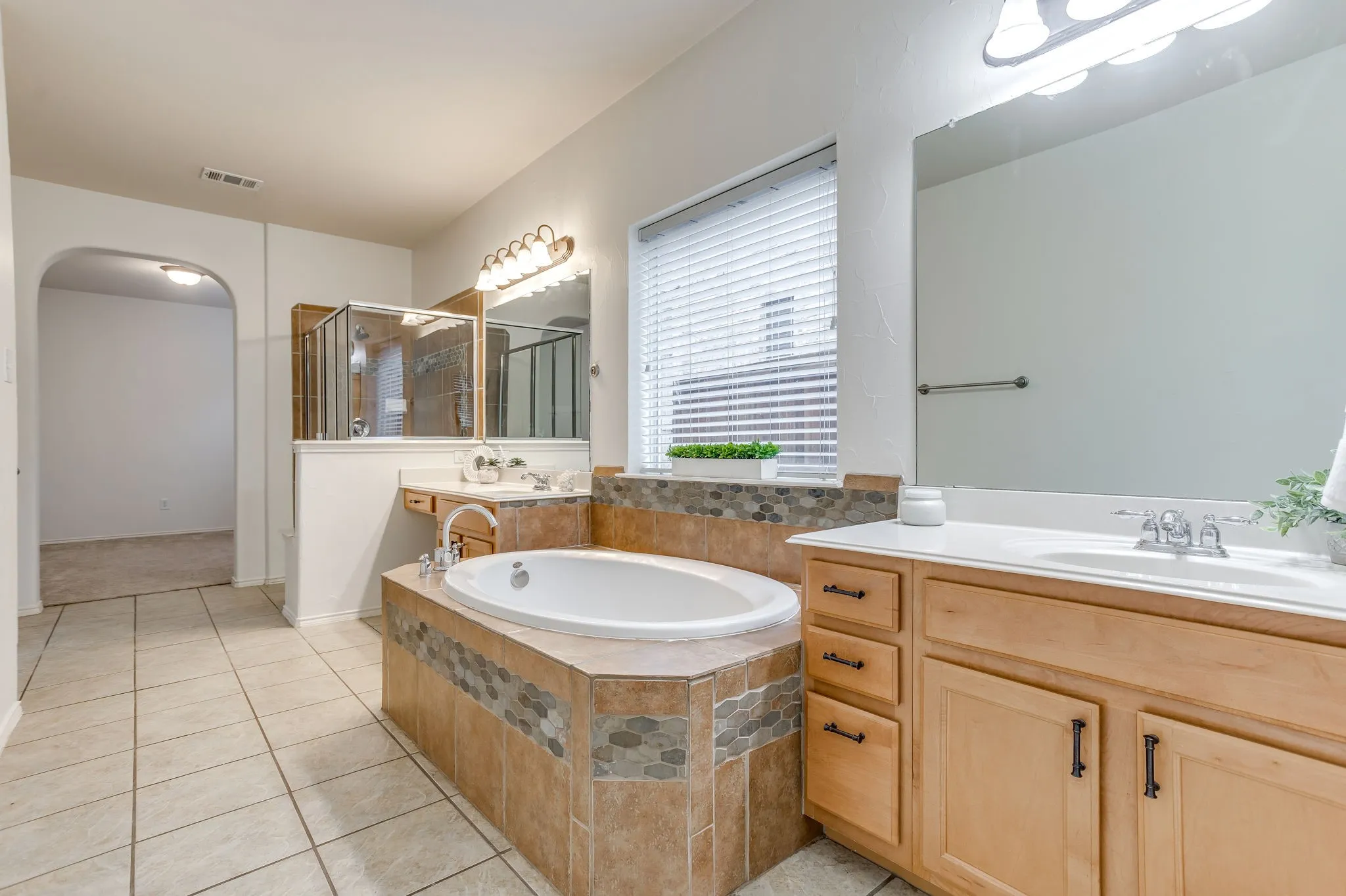 Tile flooring, built-in soaking tub with decorative tile surround, separate shower enclosure with glass doors, dual vanity sinks with wood cabinetry, and a large window with blinds