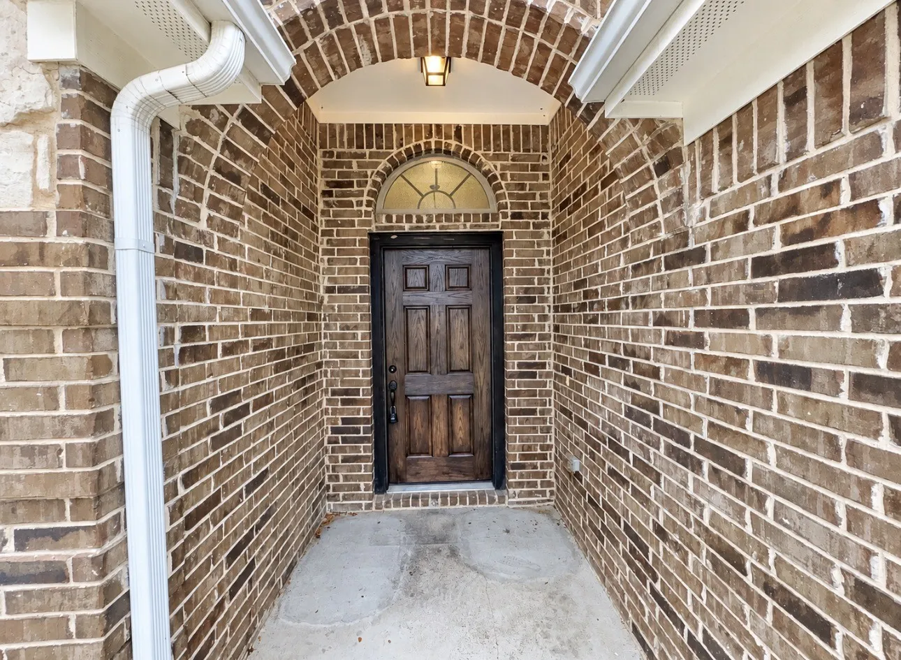 Brick exterior with an arched entryway, detailed wood-paneled door, transom window, and exterior lighting fixture