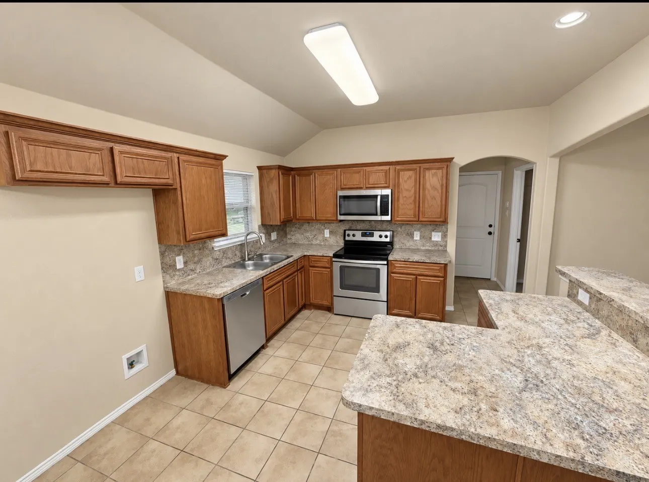Kitchen featuring wood-finish cabinetry, stainless steel appliances, speckled countertops, tile flooring, and a vaulted ceiling