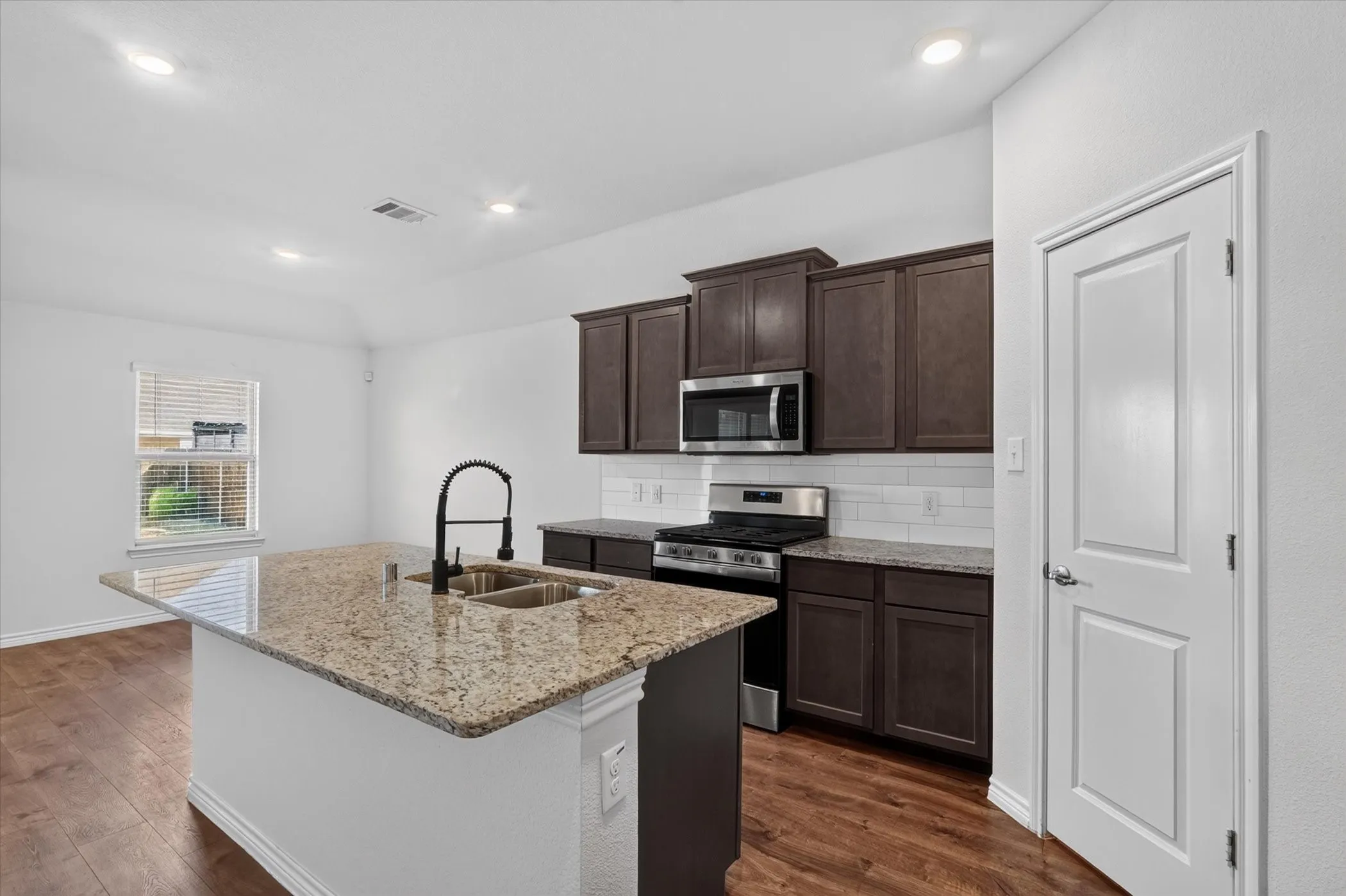 Kitchen featuring an island with granite countertops, a stainless steel double basin sink with a matte black gooseneck faucet, dark wood-finish cabinetry, and stainless steel appliances
