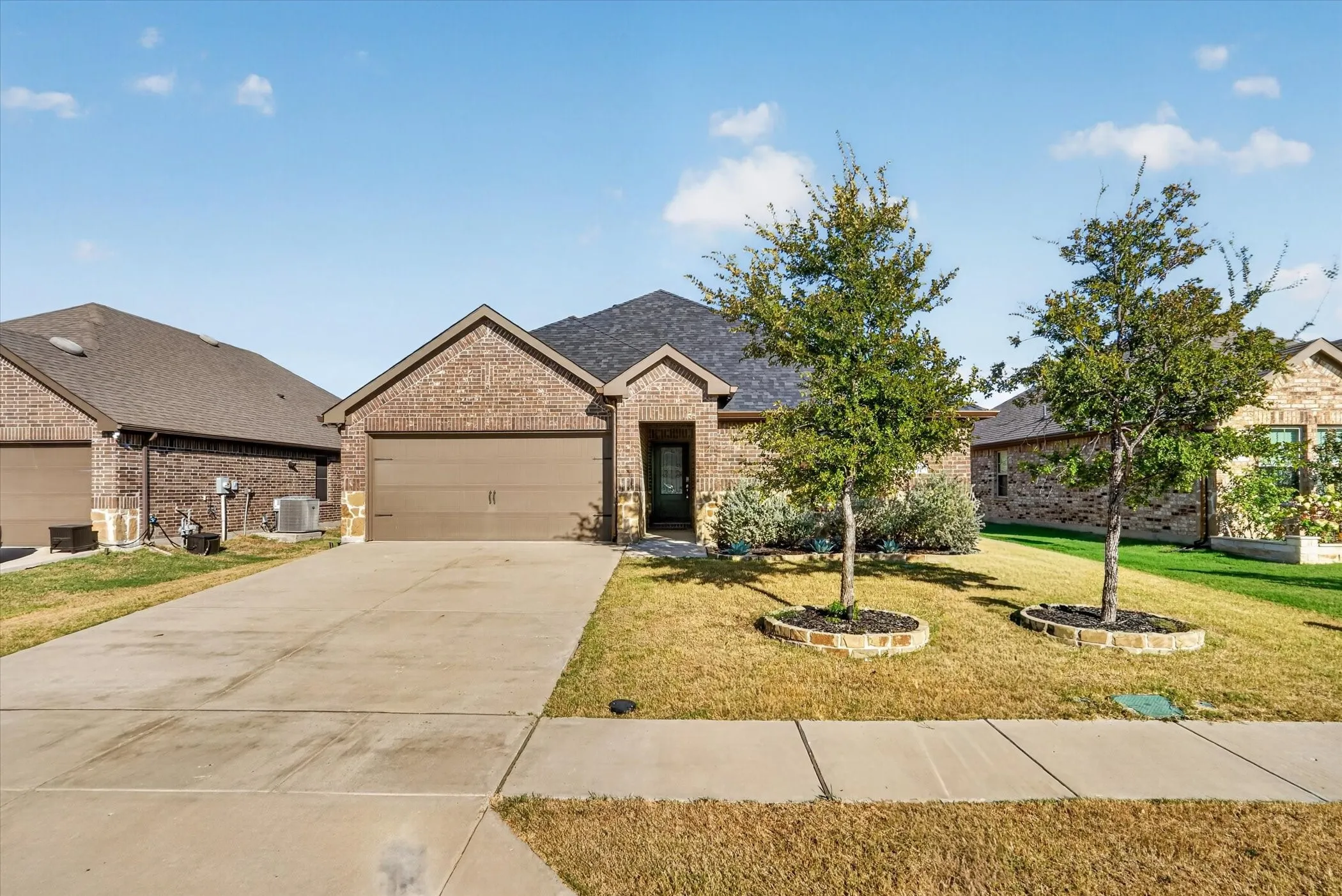 Brick exterior, asphalt shingle roof, attached two-car garage, concrete driveway, and front lawn with mature trees