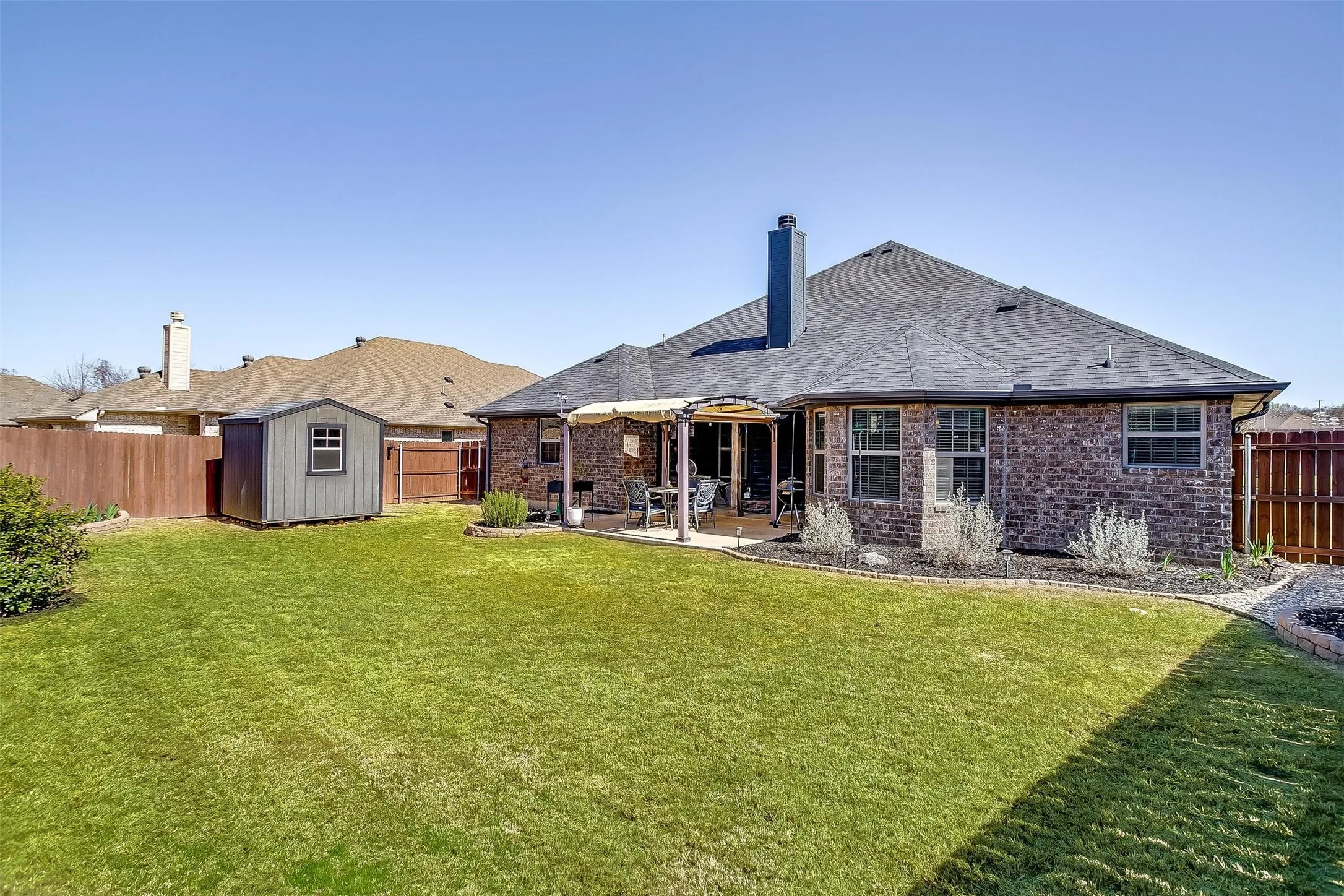 Brick exterior with multiple windows, covered patio, fenced yard, storage shed, and landscaped garden beds