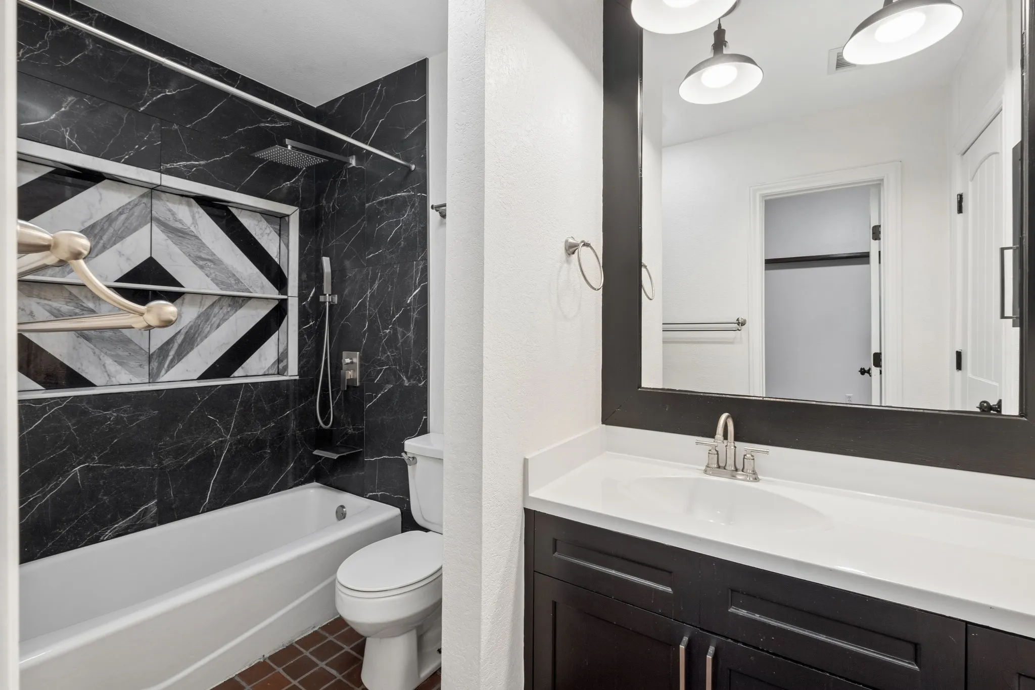 Bathtub with black marble-finish tile surround, built-in shower niche with geometric tile, black vanity with white countertop, and rectangular mirror with industrial-style sconce lighting