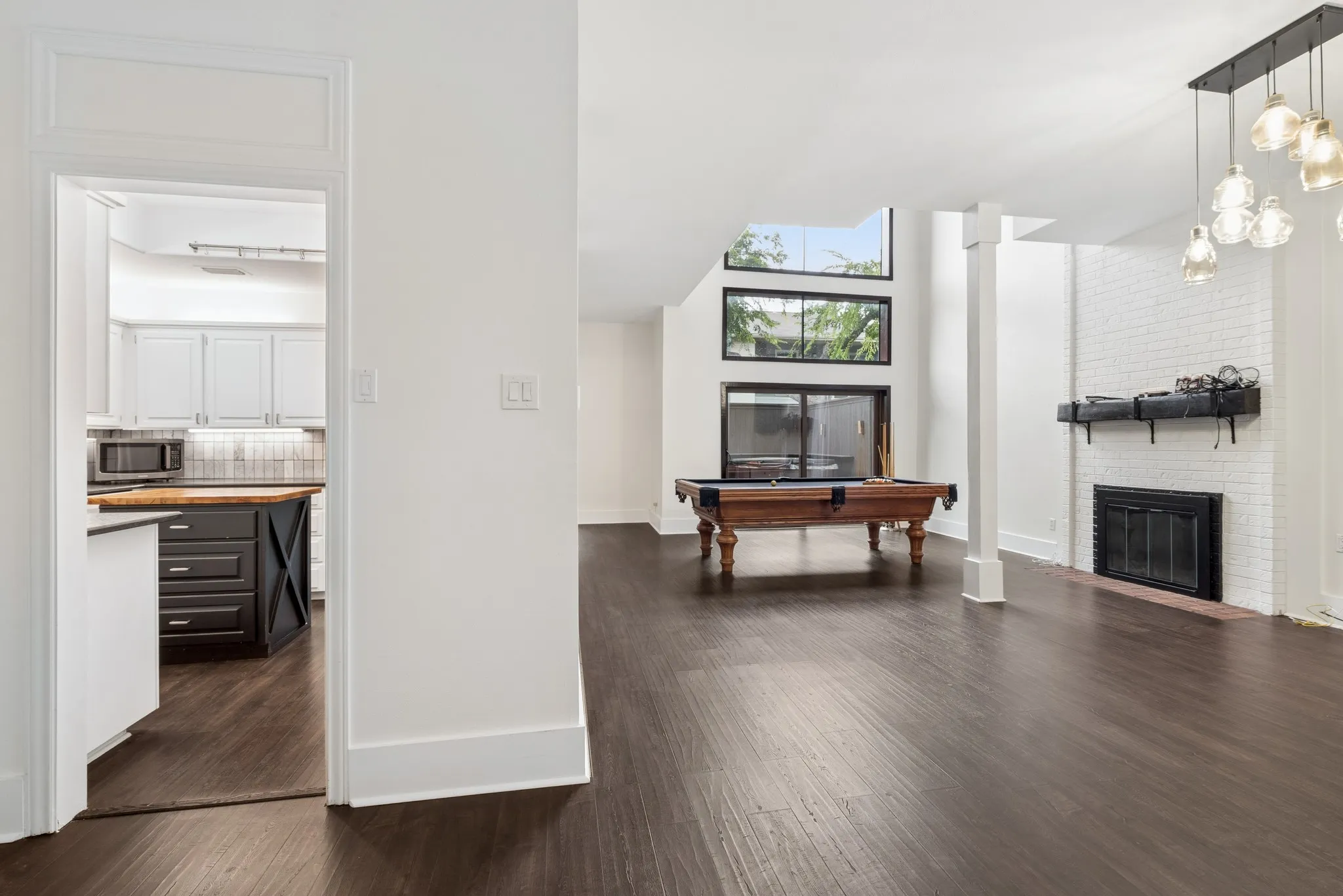 Spacious room with dark wood-finish flooring, a white brick fireplace, multiple large windows, and an attached hanging light fixture