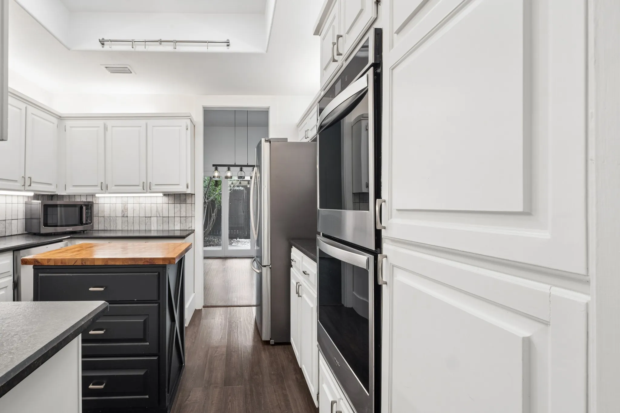 White shaker-style cabinetry, dark wood-finish flooring, stainless steel double wall ovens, butcher block island countertop, and dark stone-finish countertops
