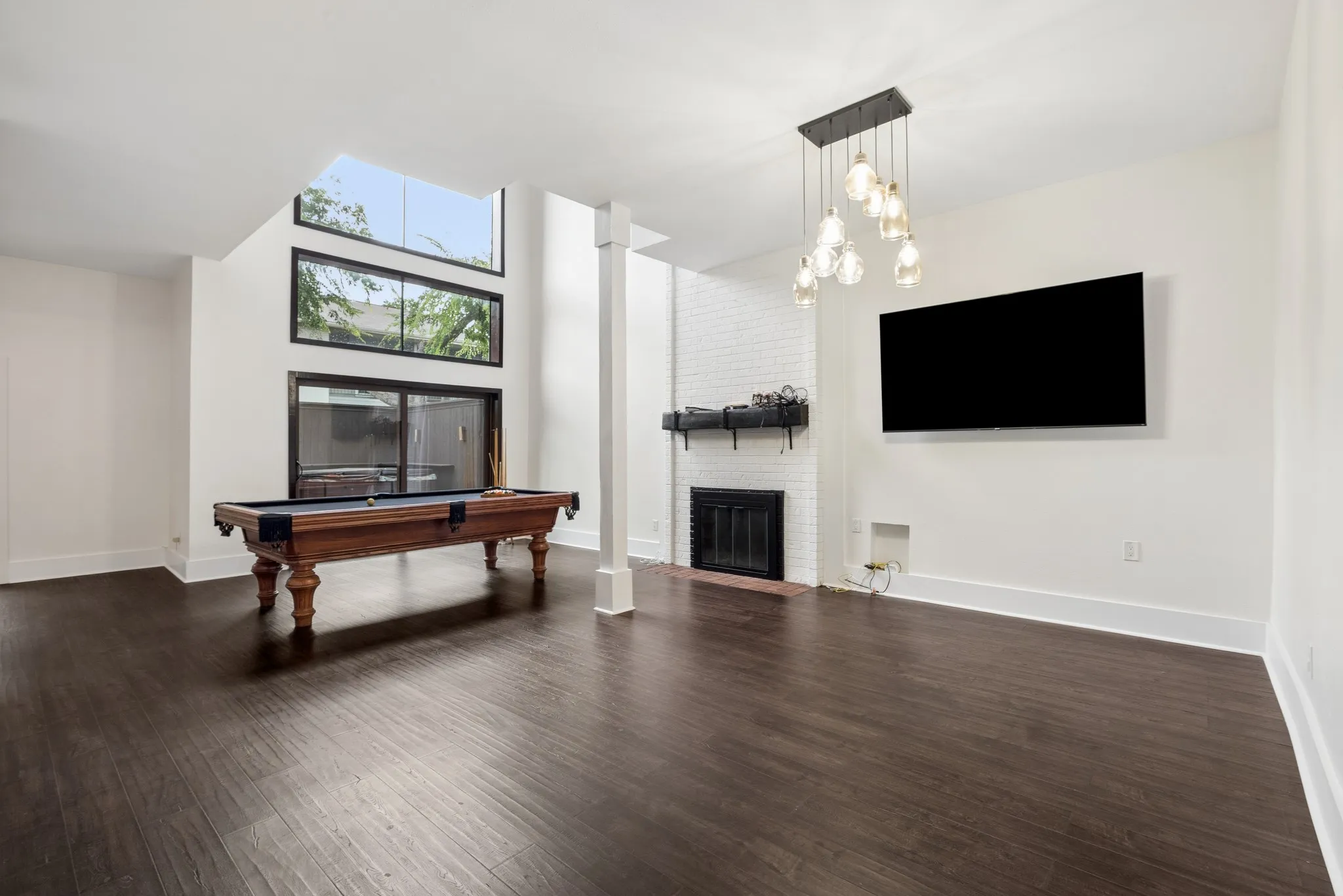 Dark wood-finish flooring, white brick fireplace with black mantel, large picture windows, recessed lighting, and a contemporary chandelier