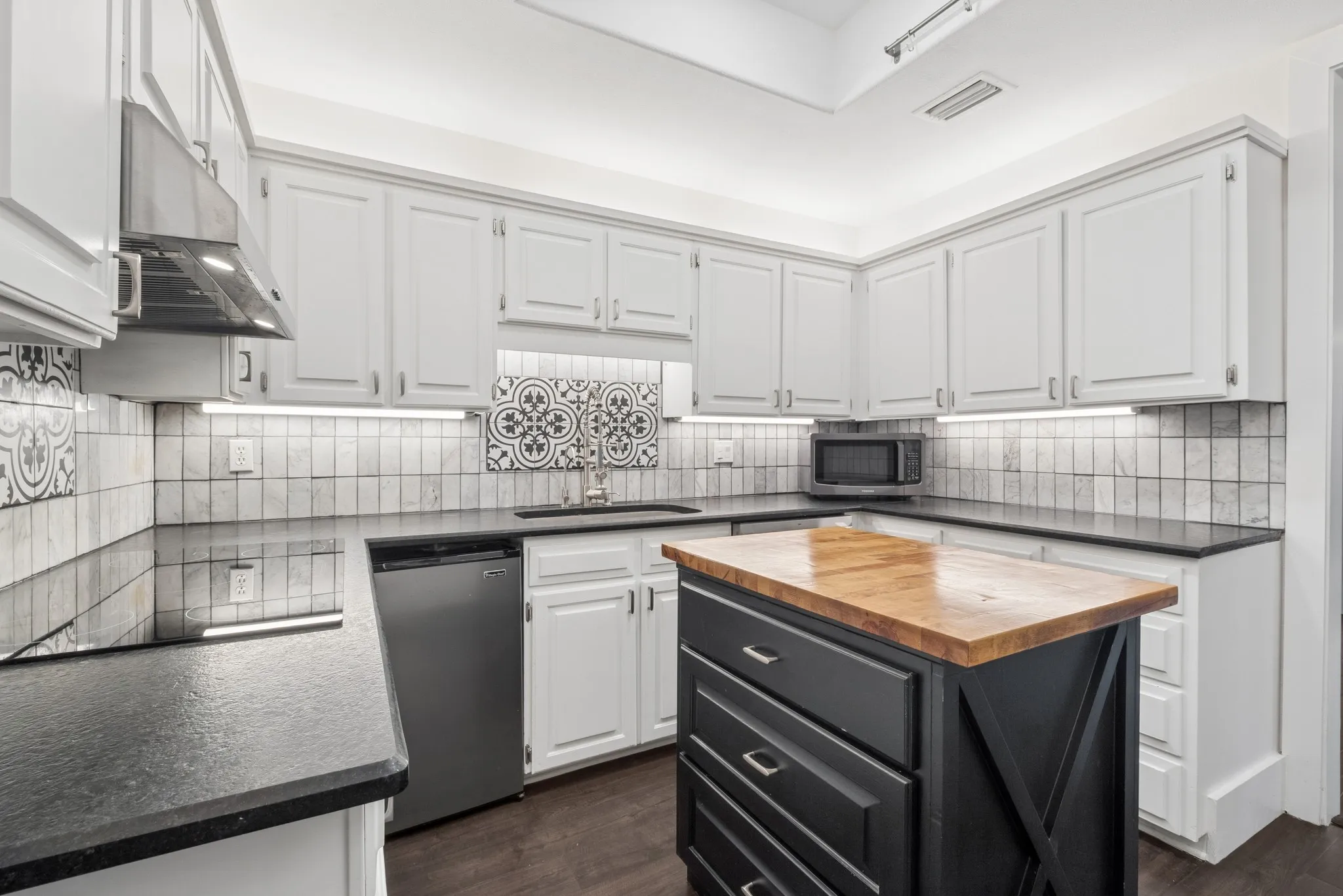 White shaker-style cabinetry with dark countertops, a custom butcher block island, stainless steel dishwasher, and a decorative tile backsplash