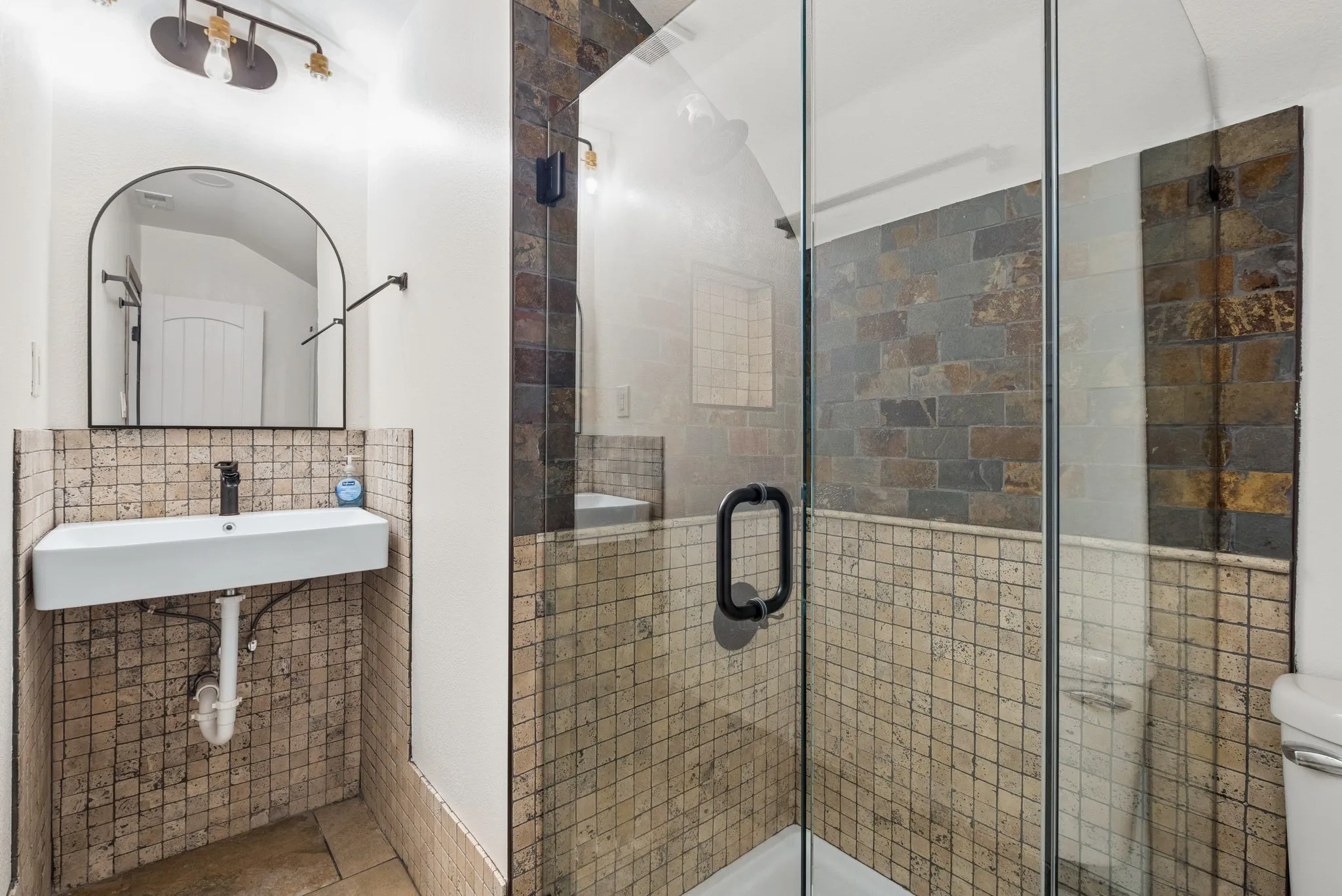 Glass-enclosed shower with stone tile surround, stone mosaic wall tiling, wall-mounted rectangular sink, and arched mirror