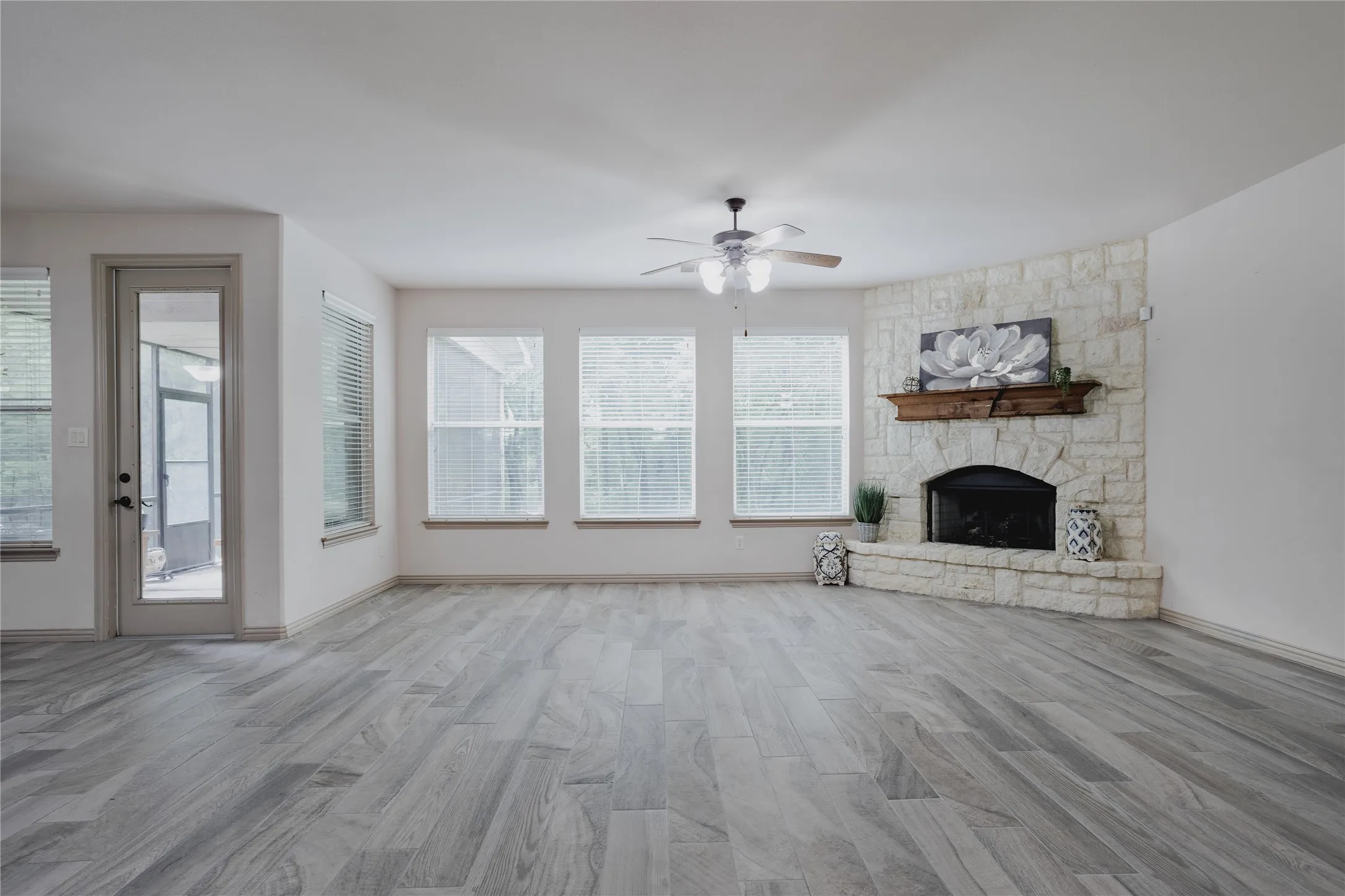 Spacious living area featuring wood-finish flooring, multiple windows, a ceiling fan, and a stone-clad corner fireplace with a wood mantel