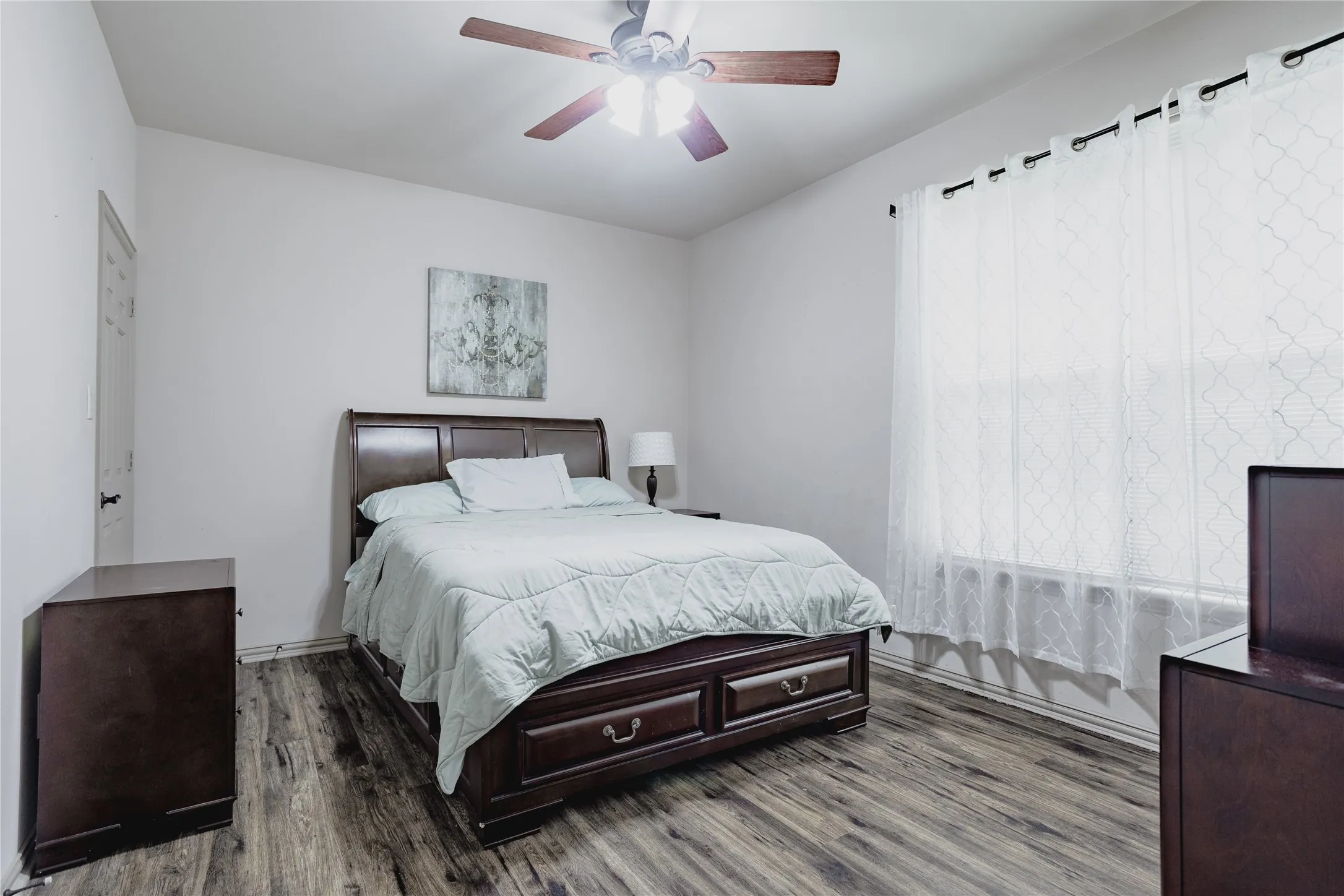 Wood-finish flooring throughout, ceiling fan with integrated lighting, and a window with sheer curtains