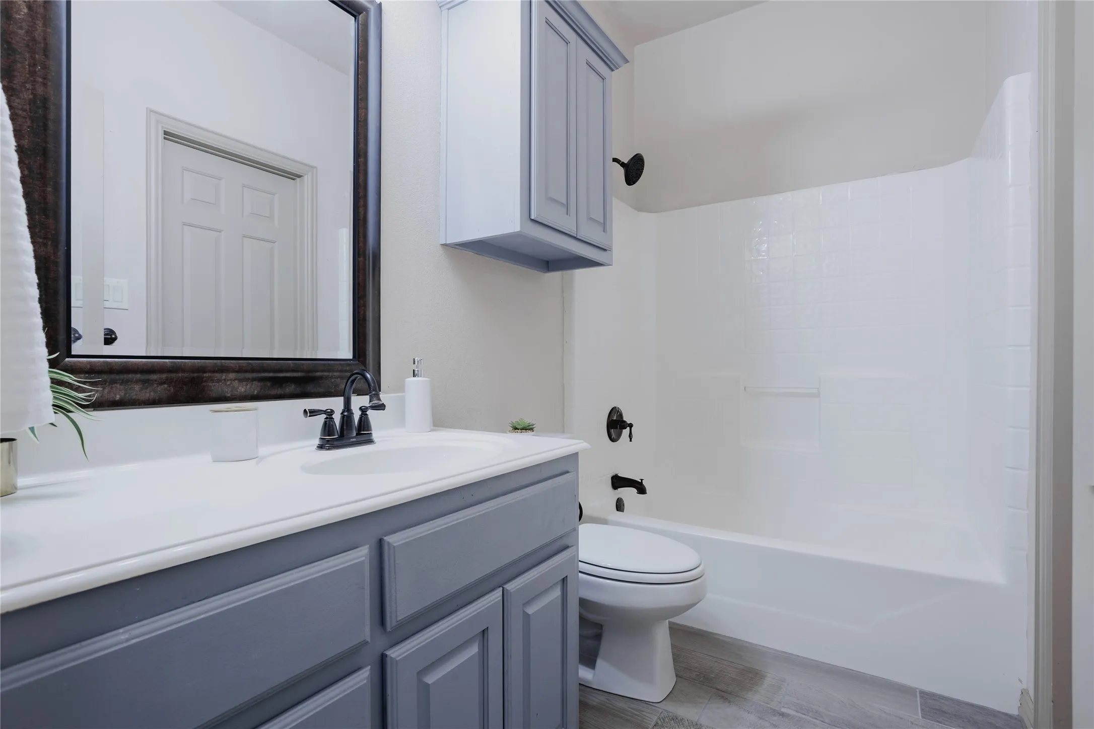 Vanity with a white countertop and gray cabinetry, dark-framed mirror, matte black fixtures, white bathtub with a shower, and gray tile flooring