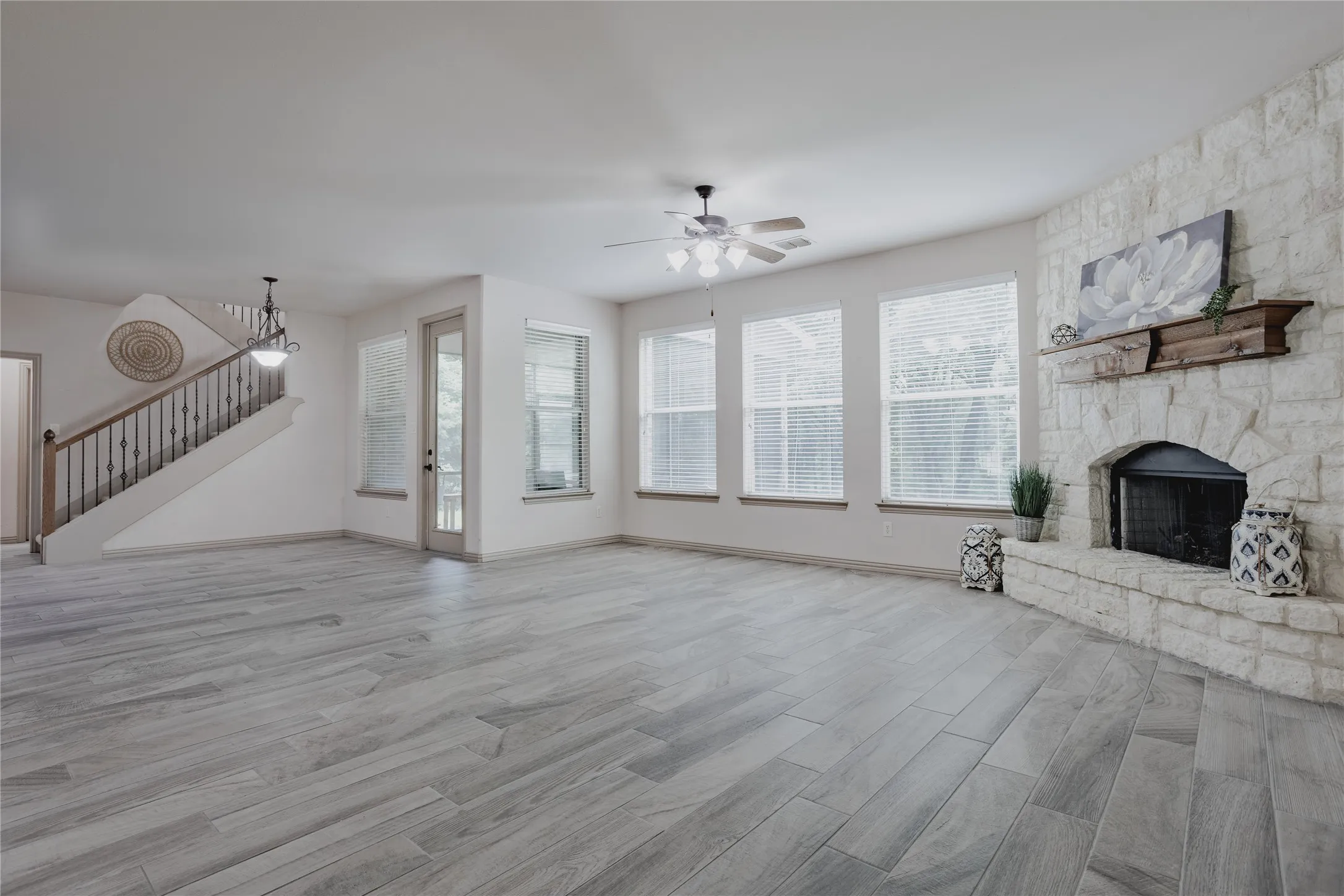 Spacious room featuring wood-finish flooring, a stone-clad fireplace with a wood mantle, multiple windows, a ceiling fan, and a staircase with wrought iron balusters