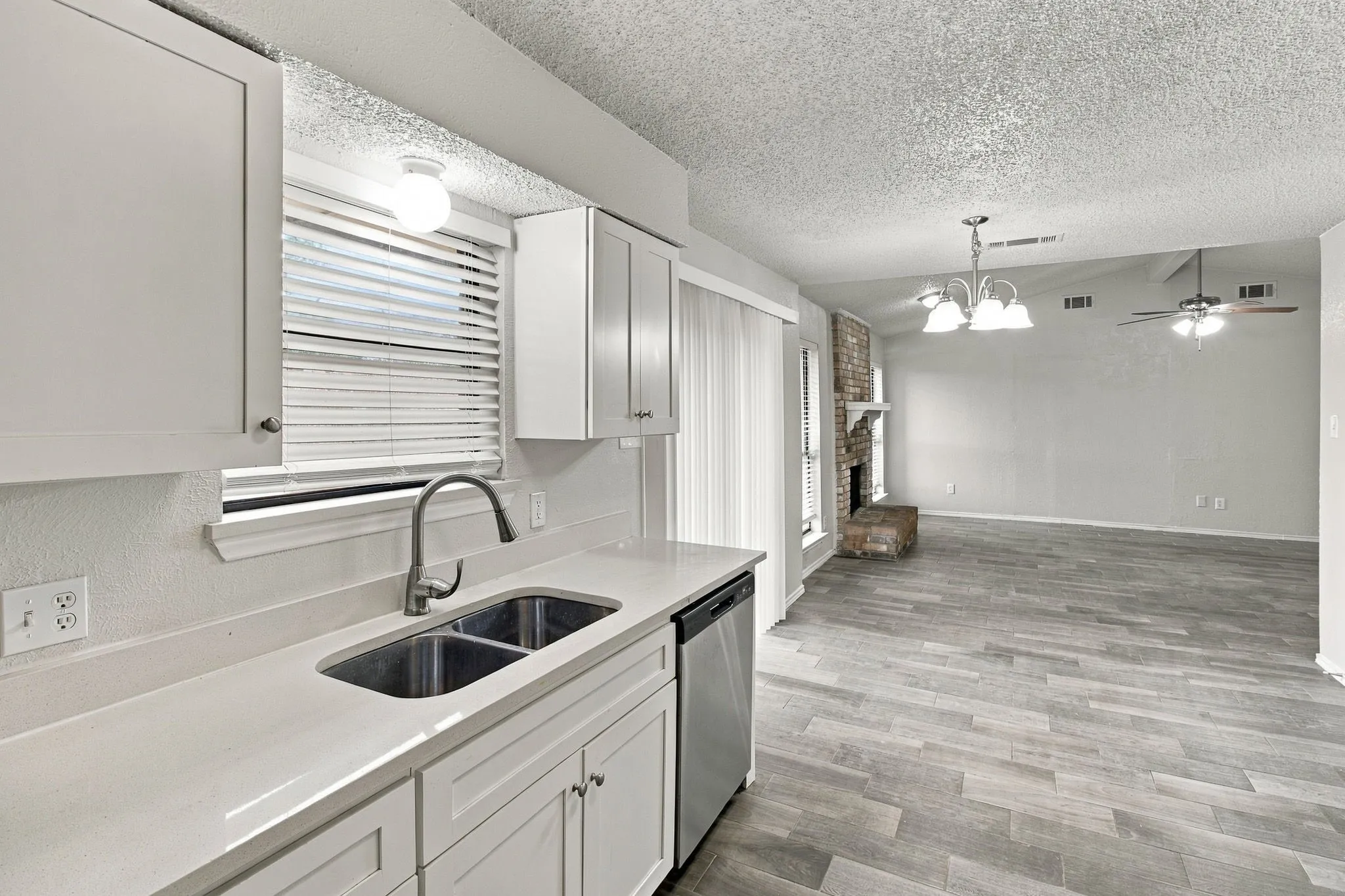 White shaker cabinetry, dual basin stainless steel sink, light-toned countertops, wood-finish flooring, and a brick-finish fireplace