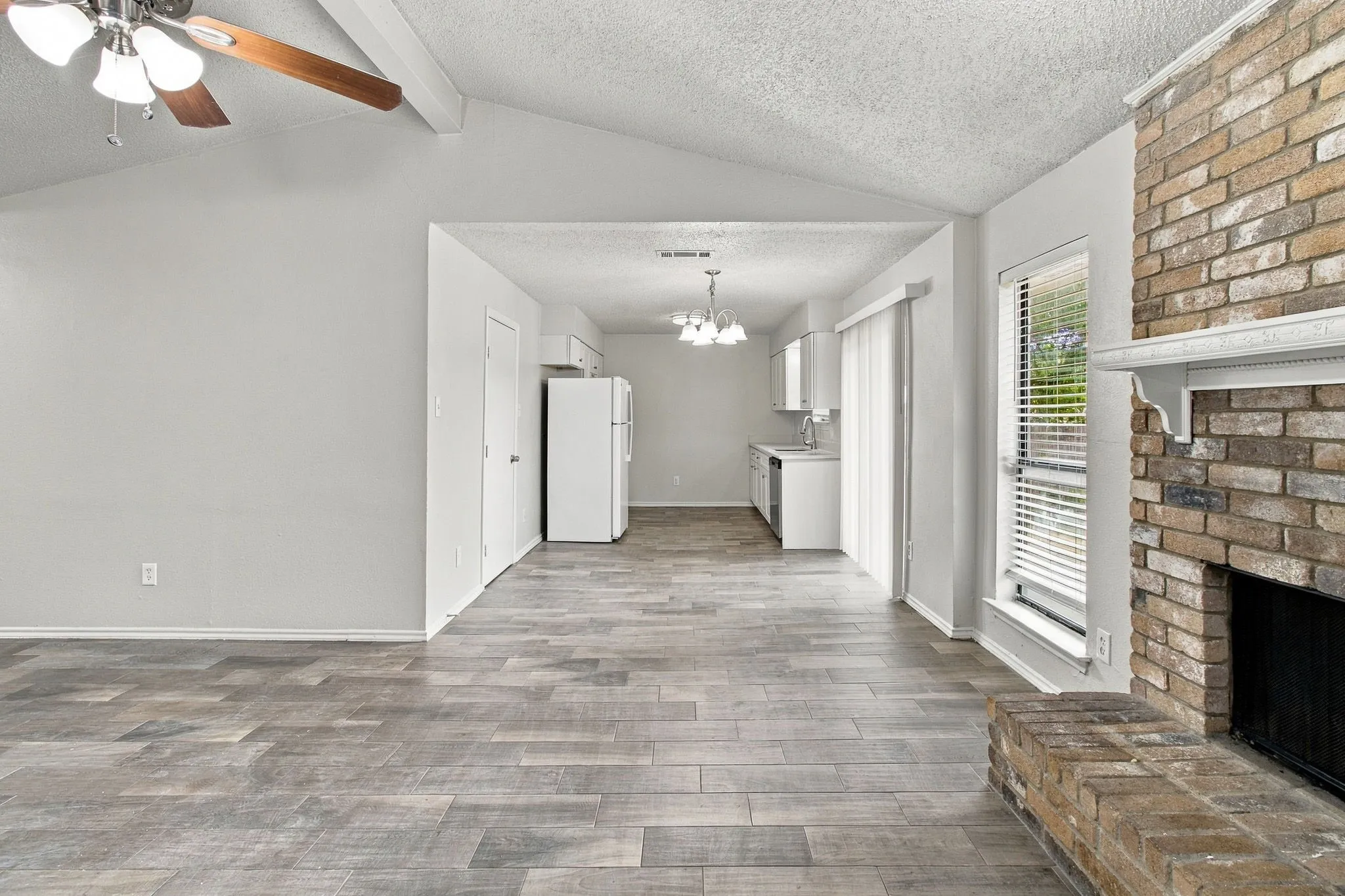 Vaulted ceiling with exposed beam and ceiling fan, brick fireplace with decorative mantel, wood-finish flooring, and kitchen with white cabinetry