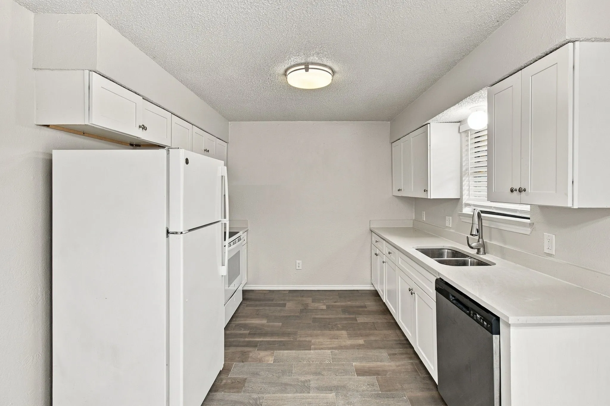 Galley kitchen featuring white cabinetry, light countertops, wood-finish flooring, a double basin sink with a gooseneck faucet, and a built-in dishwasher