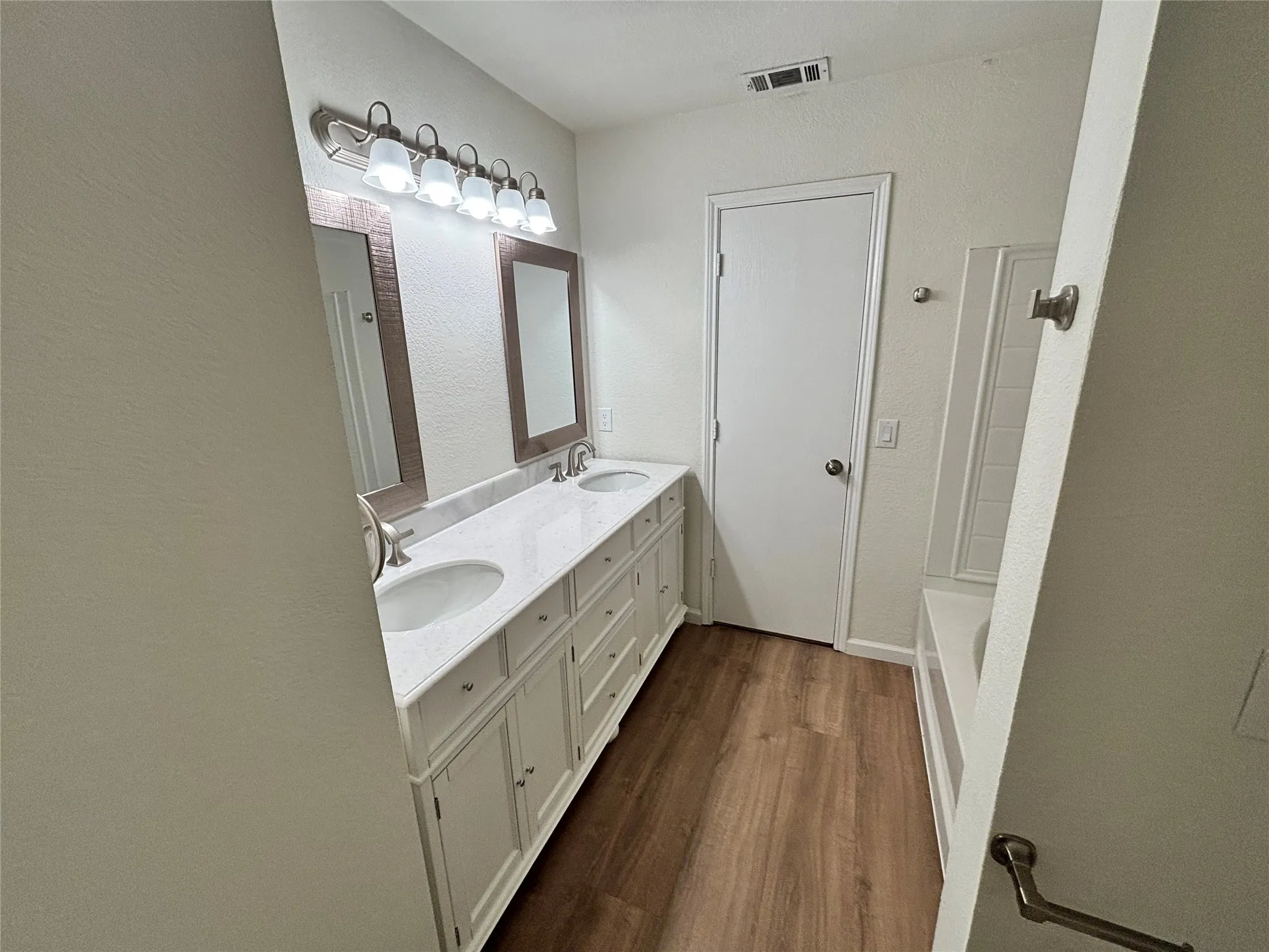 Dual vanity with integrated sinks, wood-finish flooring, two framed mirrors, brushed nickel light fixture, and a built-in tub with tile surround