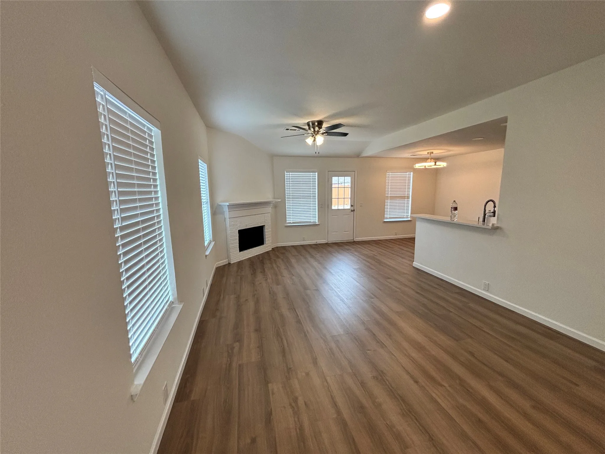 Expansive open-concept living space with wood-finish flooring, a white mantel fireplace, and a ceiling fan with integrated lighting