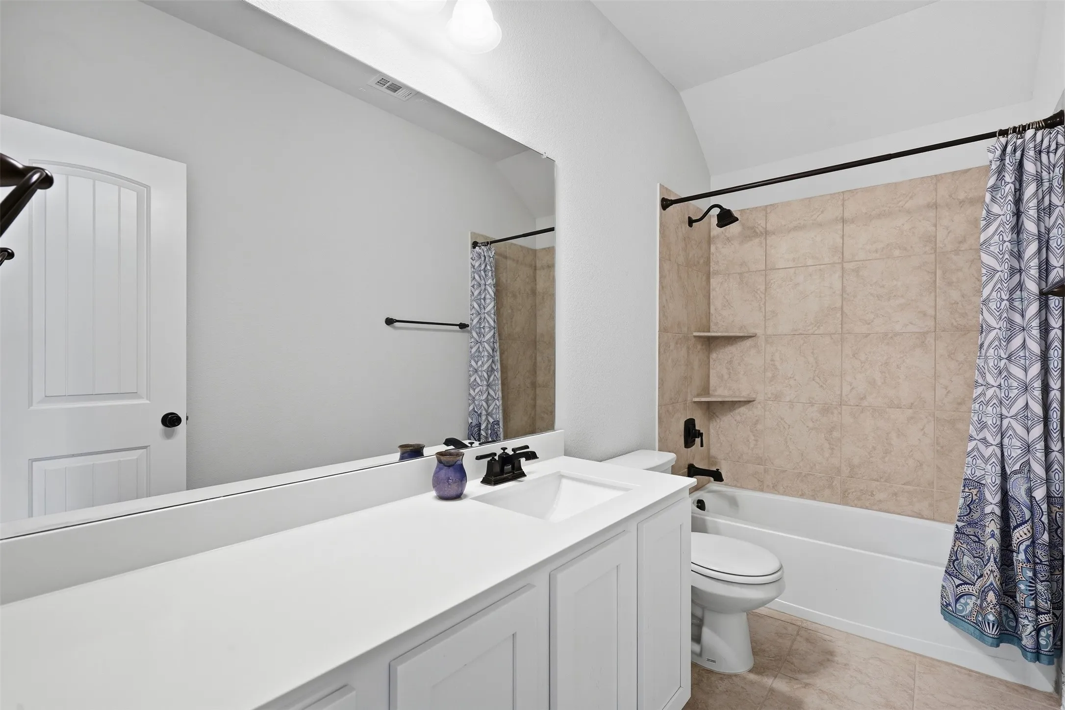 White vanity with an integrated sink, a large wall-mounted mirror, a tub and shower combination with tan tile surround, and black fixtures