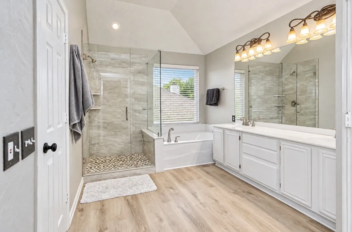 Bathroom featuring a glass-enclosed shower with pebble flooring and tiled walls, a separate built-in soaking tub, an extended vanity with dual sinks, and light wood-finish flooring