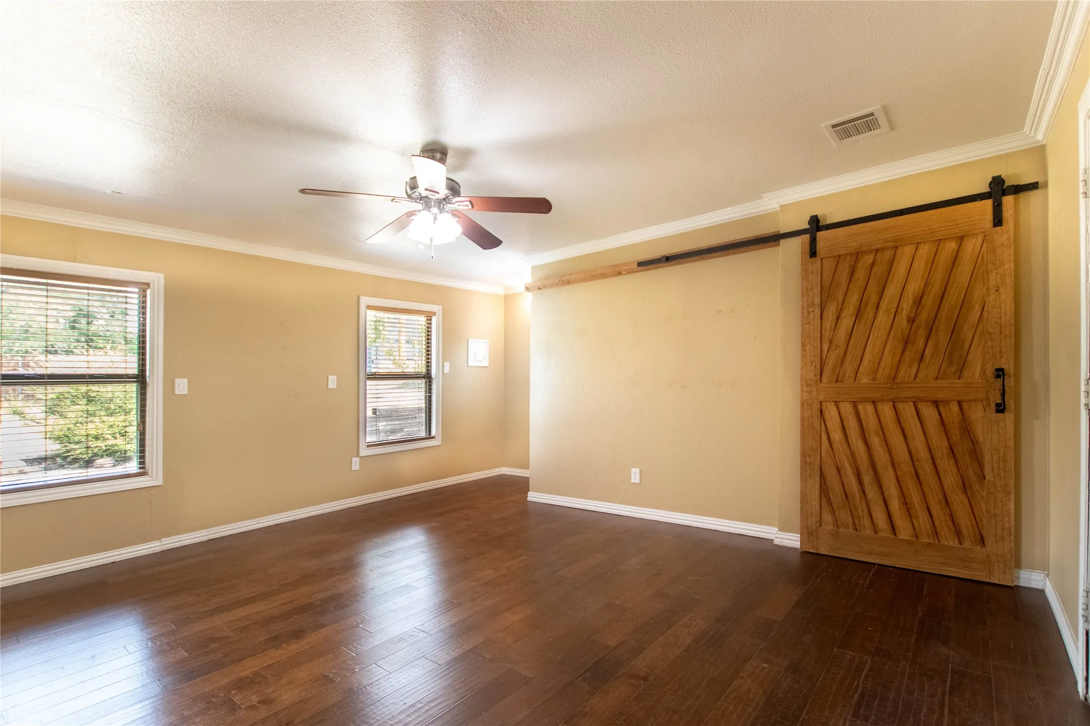 Dark wood-finish flooring, two windows with blinds, crown molding, ceiling fan with integrated lighting, and a wood barn door with a black track
