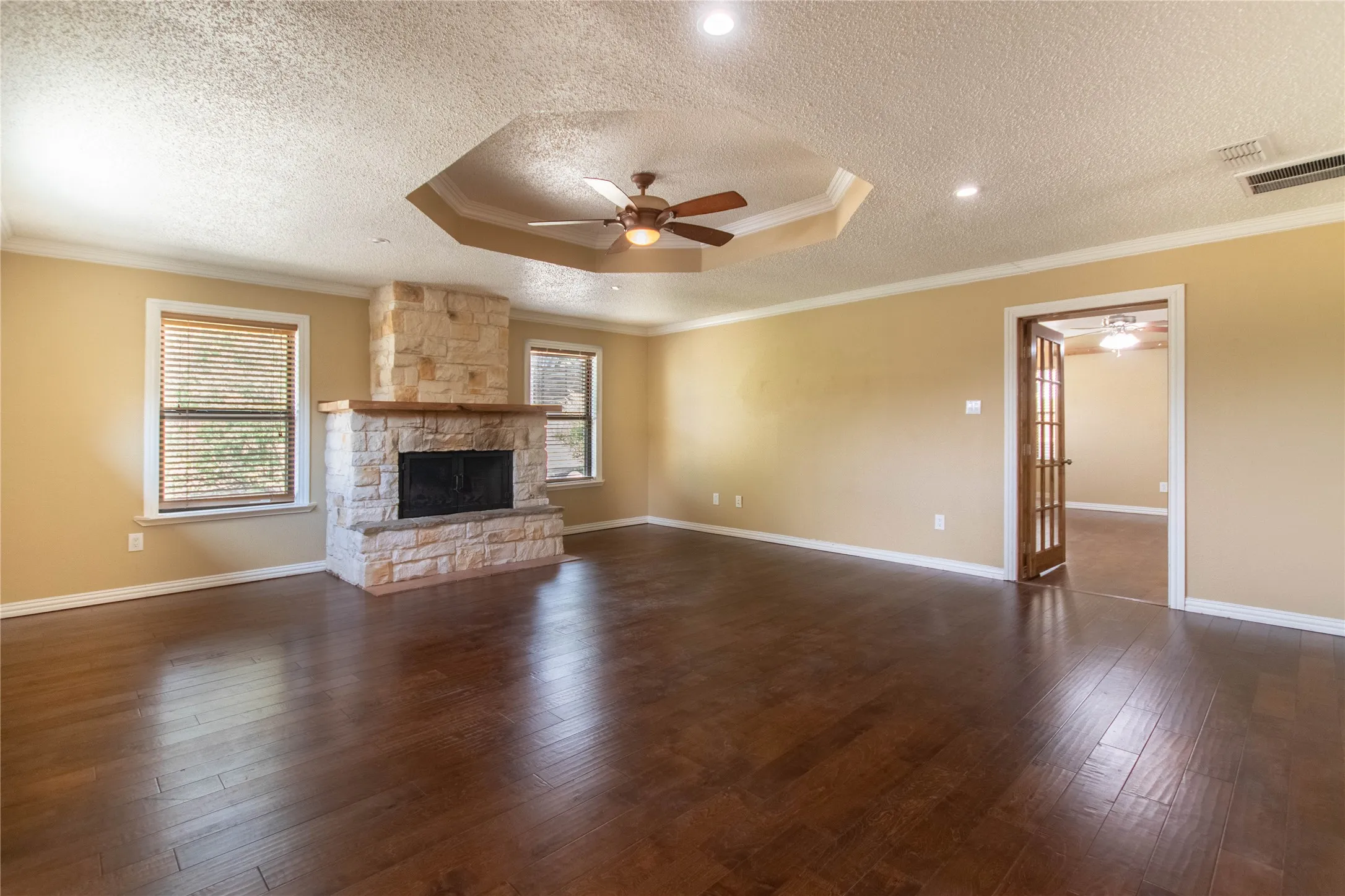 Wood-finish flooring, stone-clad fireplace with wood mantle, recessed lighting, ceiling fan, and tray ceiling