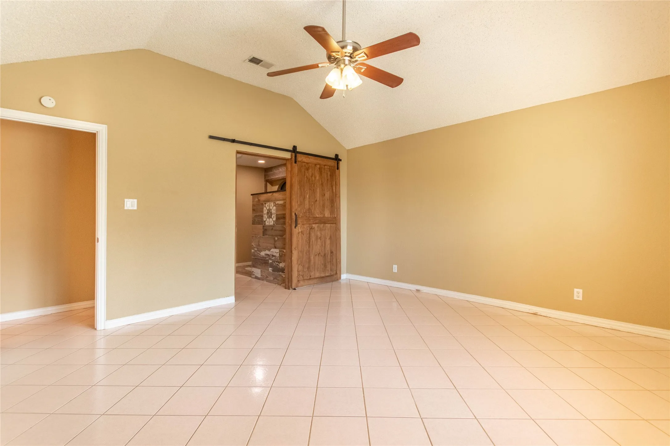 Vaulted ceiling, ceiling fan with light fixture, light-toned floor tile, wood barn door, and stone accent wall