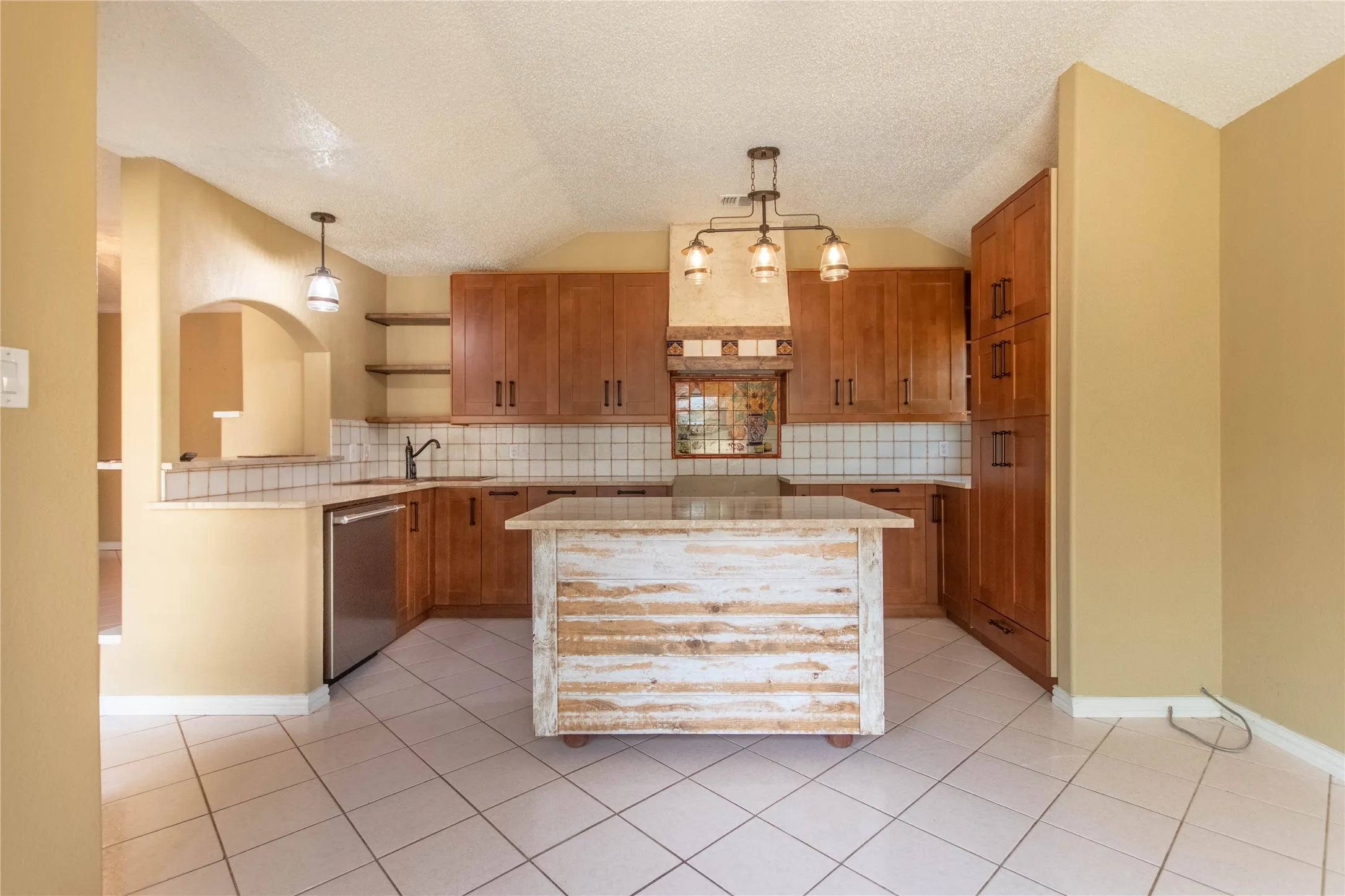 Kitchen featuring wood cabinetry, tile flooring, a central island, stainless steel dishwasher, and a decorative light fixture
