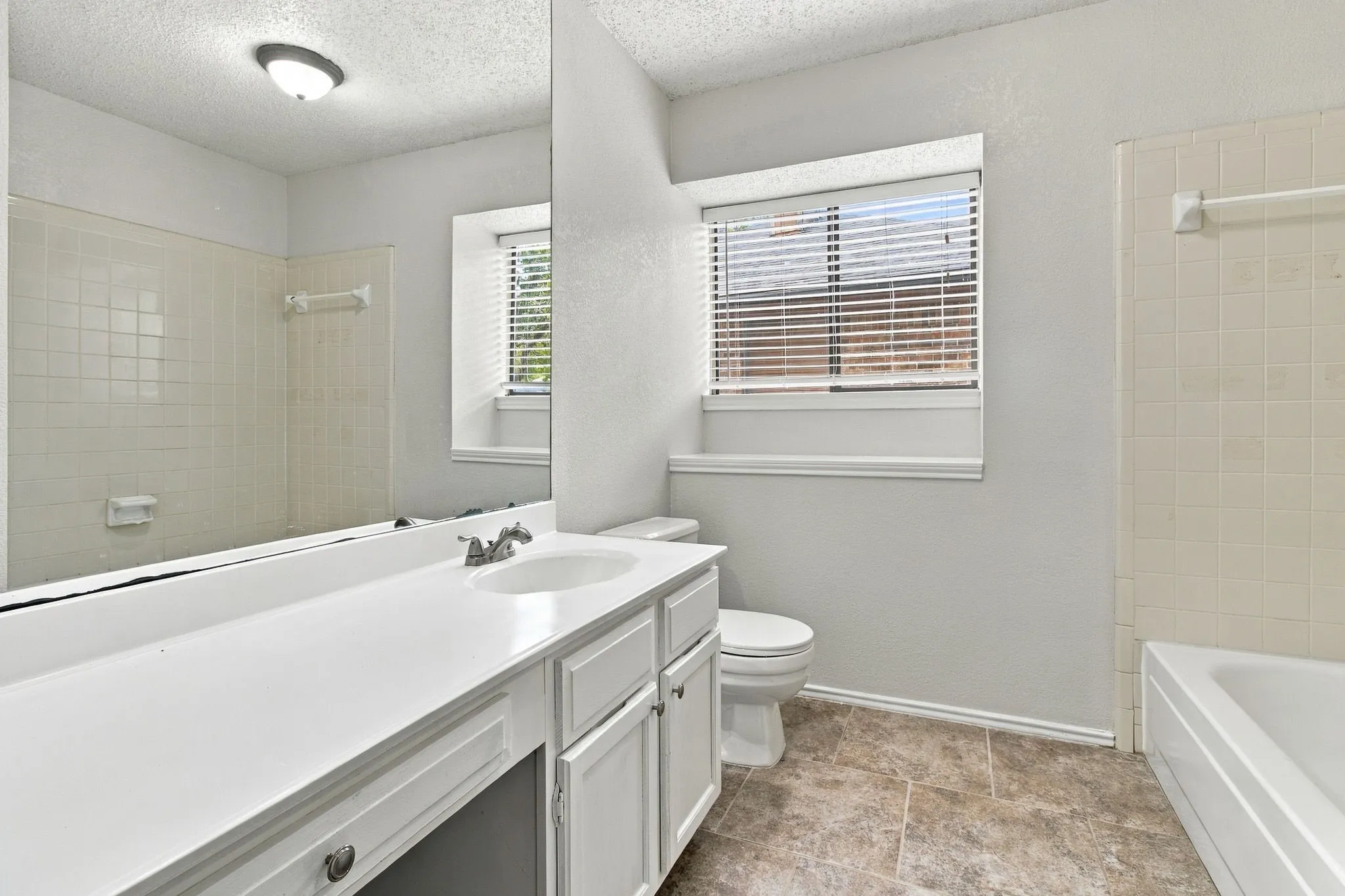 Bathroom featuring a vanity with integrated sink, a large mirror, a window with blinds, a toilet, and a shower/tub combination with tiled surround