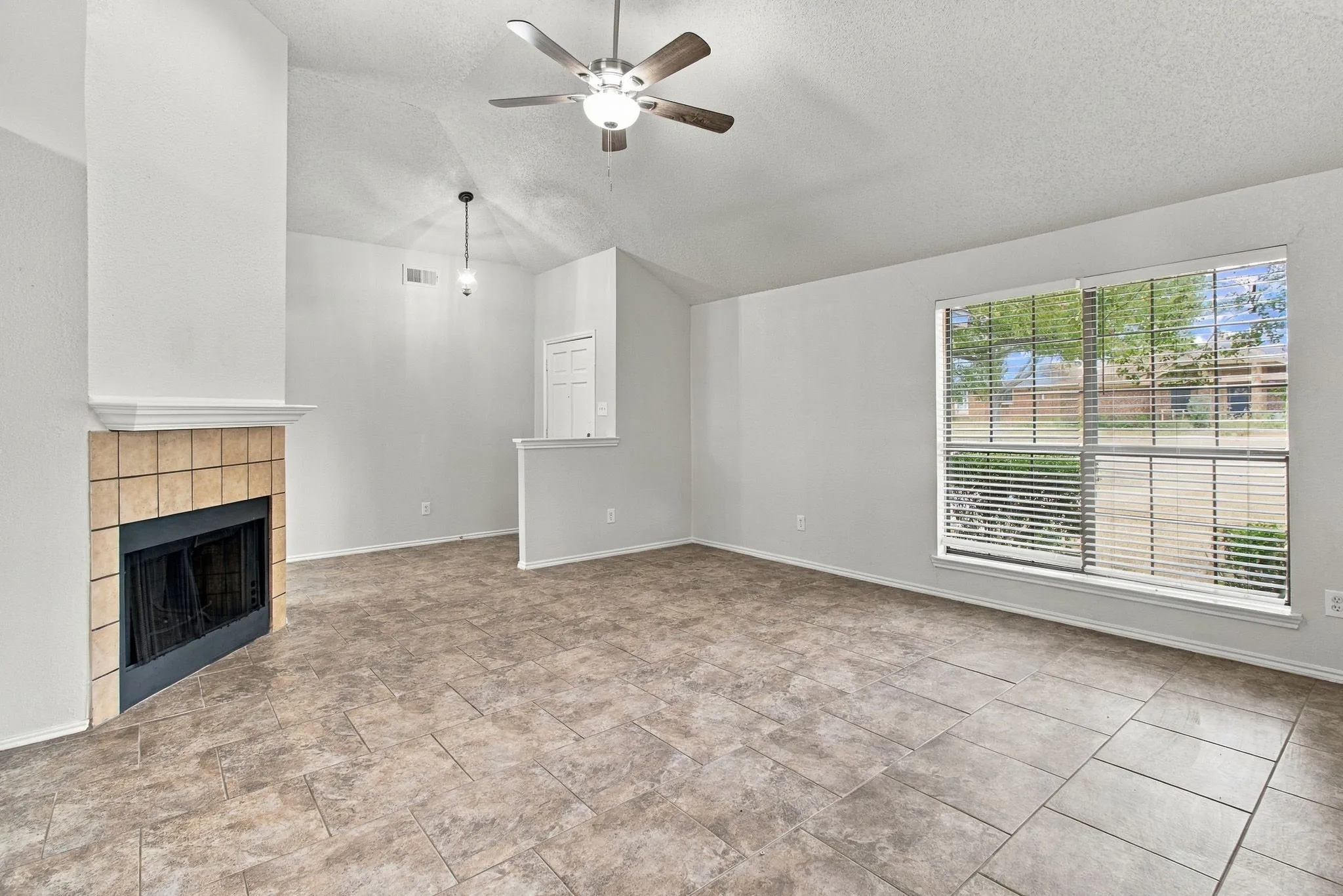 Spacious room featuring vaulted ceilings, a tiled fireplace with mantel, a ceiling fan, and large windows with horizontal blinds