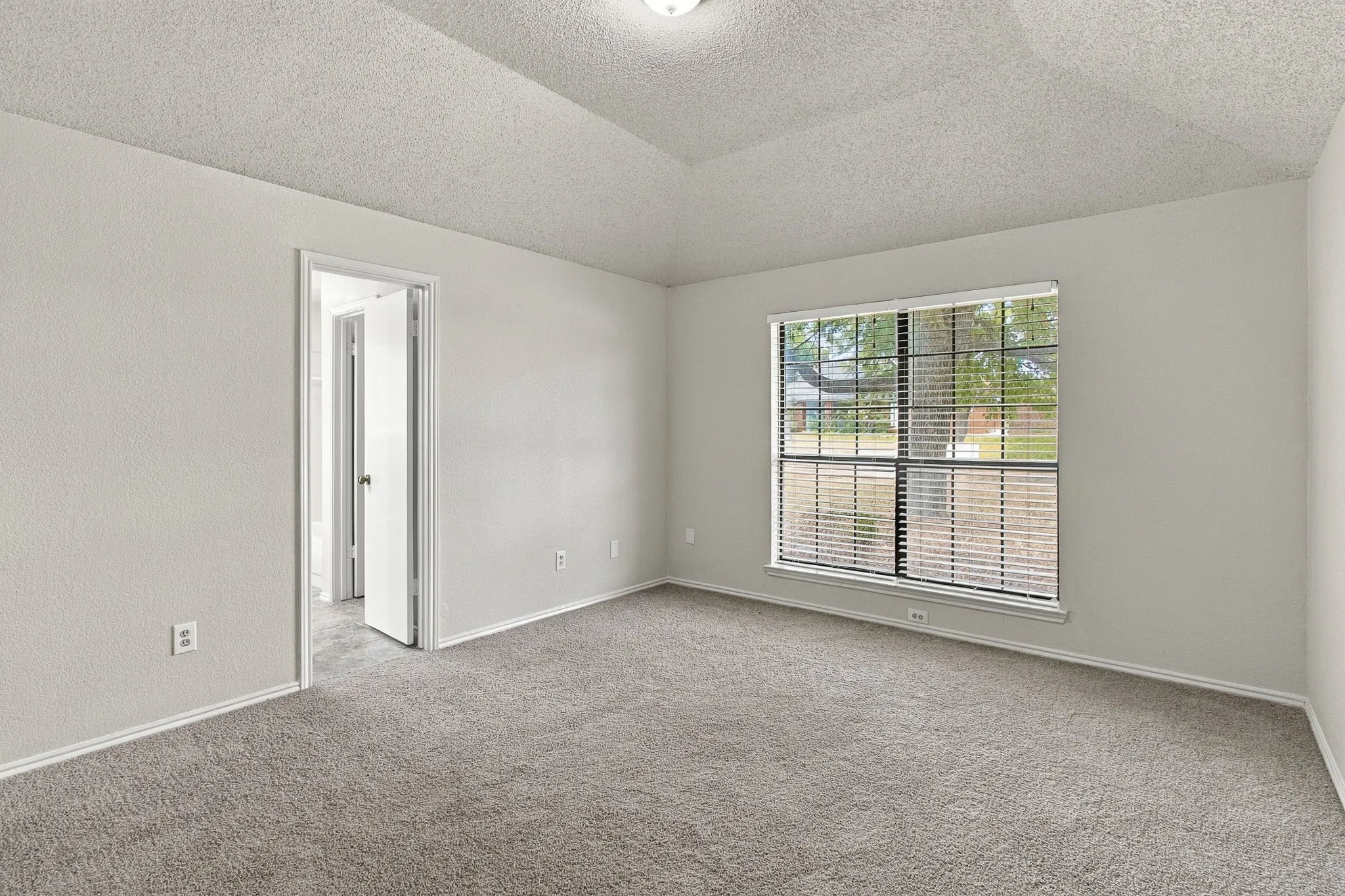 Vaulted ceiling, wall-to-wall carpeting, single window with blinds, light-toned wall paint, and white trim throughout