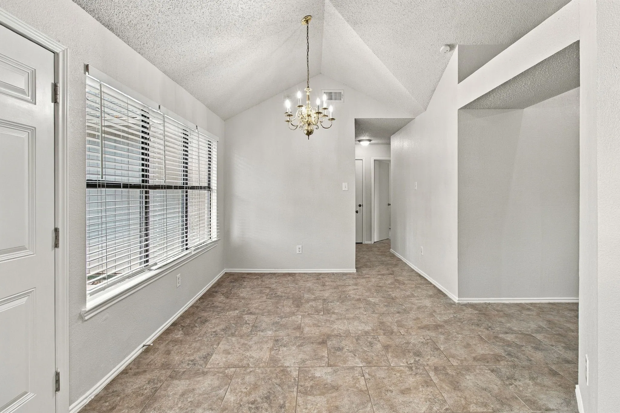 Vaulted ceiling with an overhead chandelier, large window with blinds, tile flooring, and a two-panel interior door