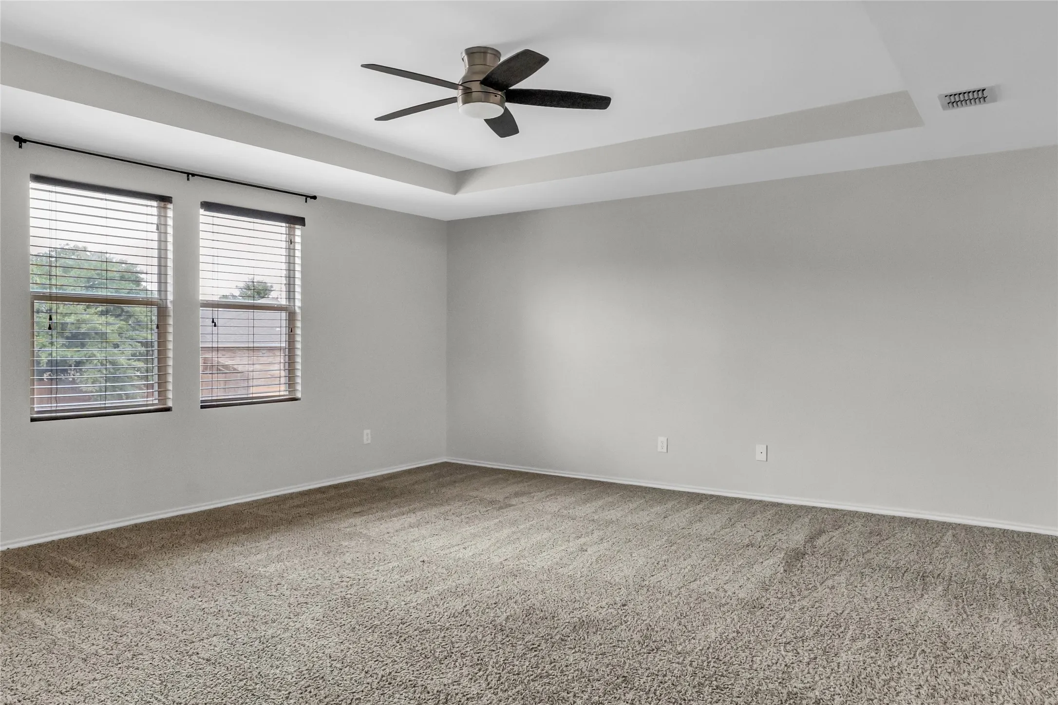 Tray ceiling with a ceiling fan, dual windows with blinds, and neutral carpeting
