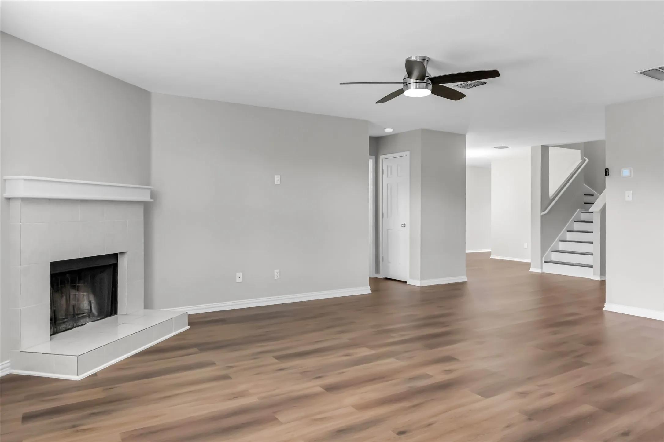 Expansive room featuring wood-finish flooring, a contemporary fireplace with a tiled hearth, a ceiling fan with integrated lighting, and a prominent staircase with white risers