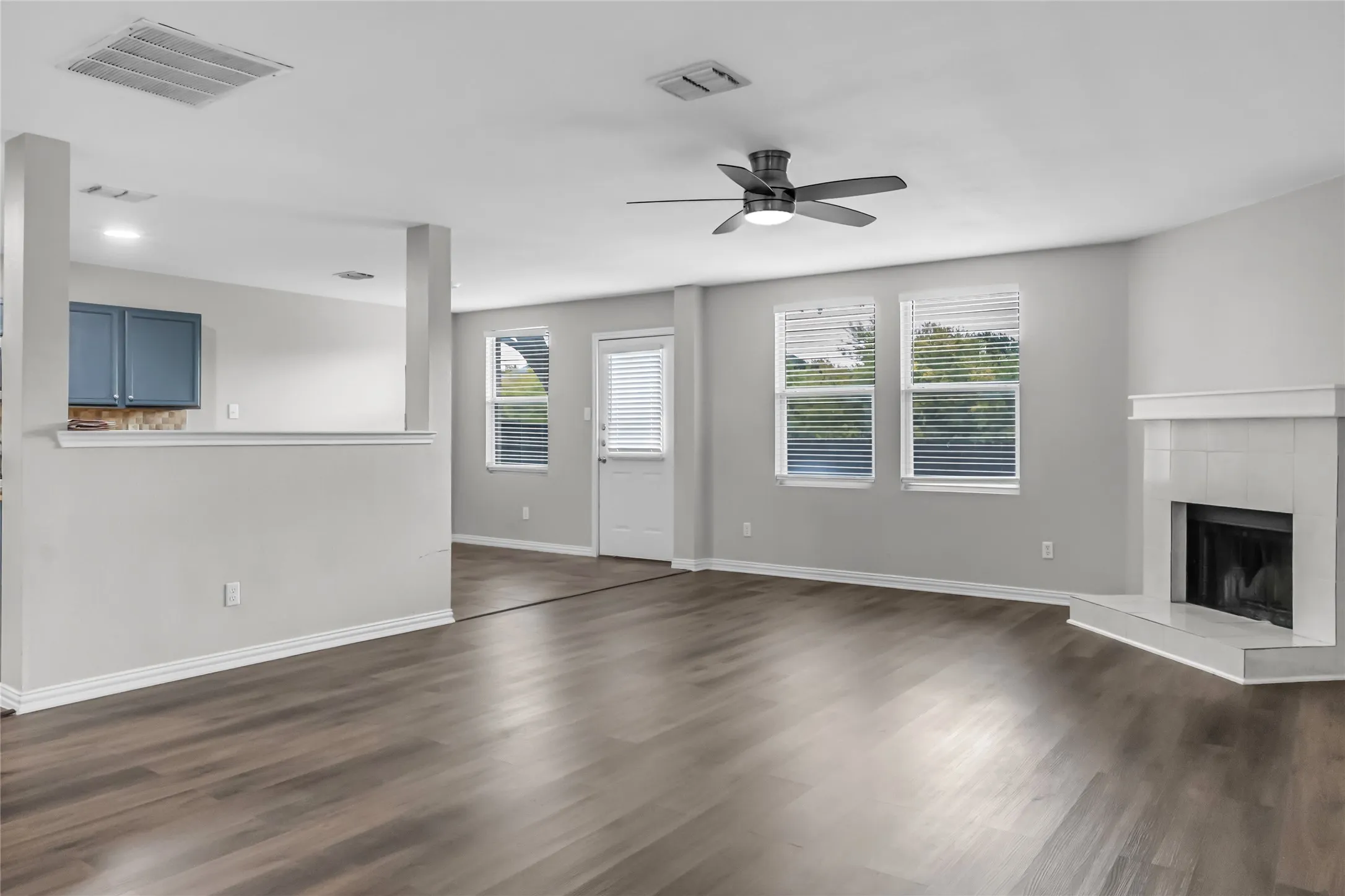 Wood-finish flooring throughout, white-painted corner fireplace, ceiling fan with integrated lighting, multiple windows with blinds, and a built-in half wall