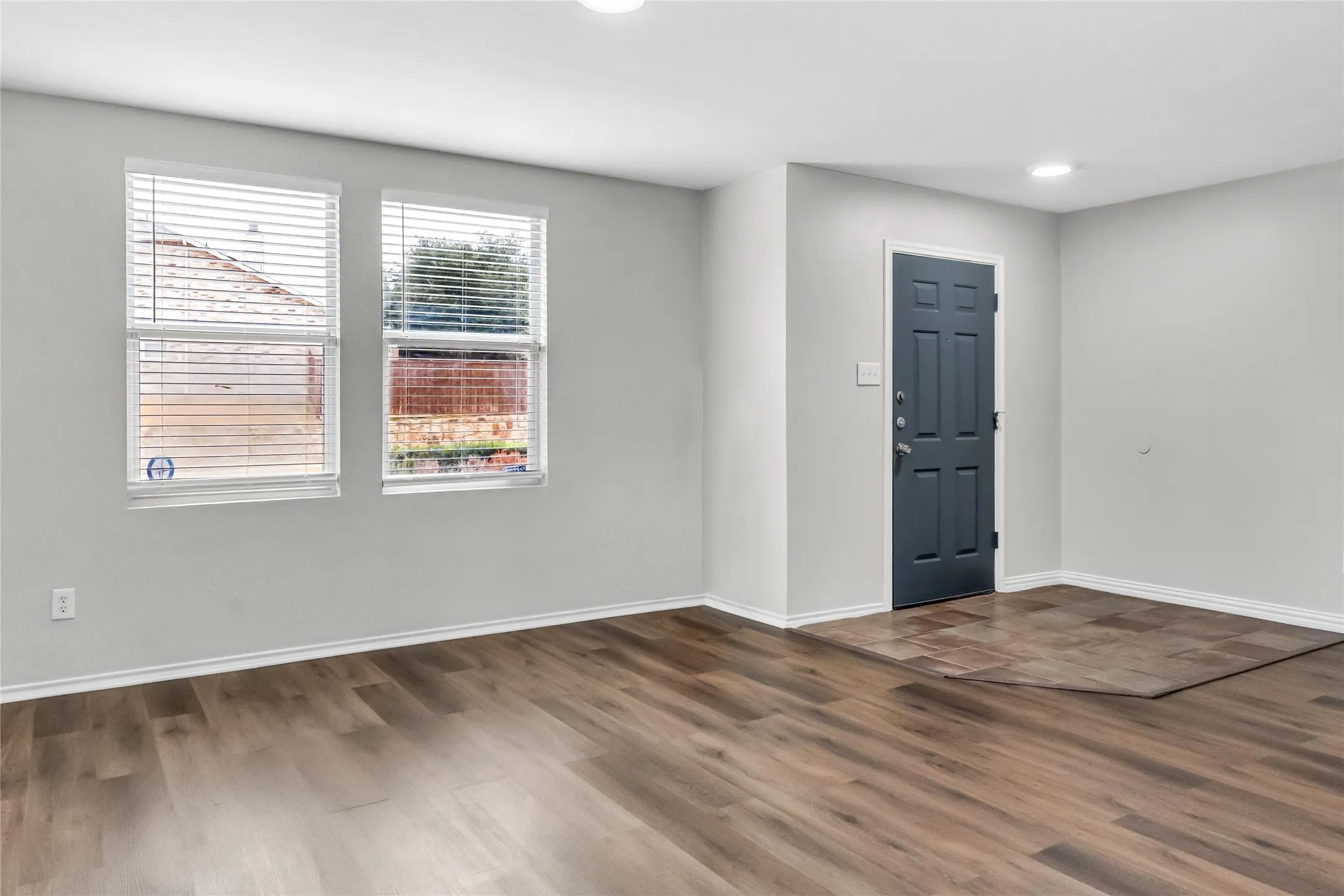 Wood-finish flooring throughout, bright walls, two windows with blinds, a dark paneled door, and recessed lighting