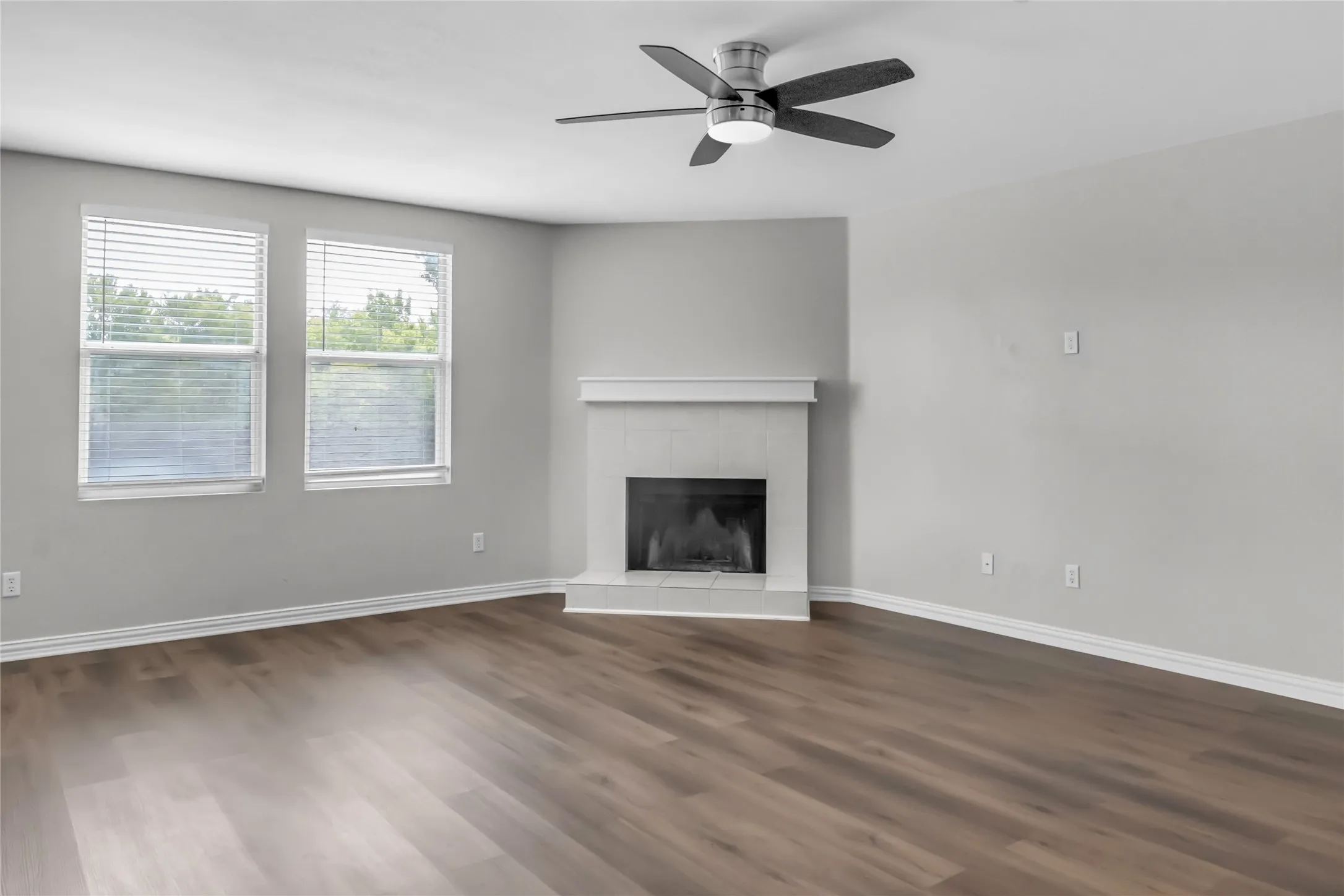 Wood-finish flooring throughout, two large windows with blinds, a corner fireplace with a white mantel, and a ceiling fan with integrated lighting